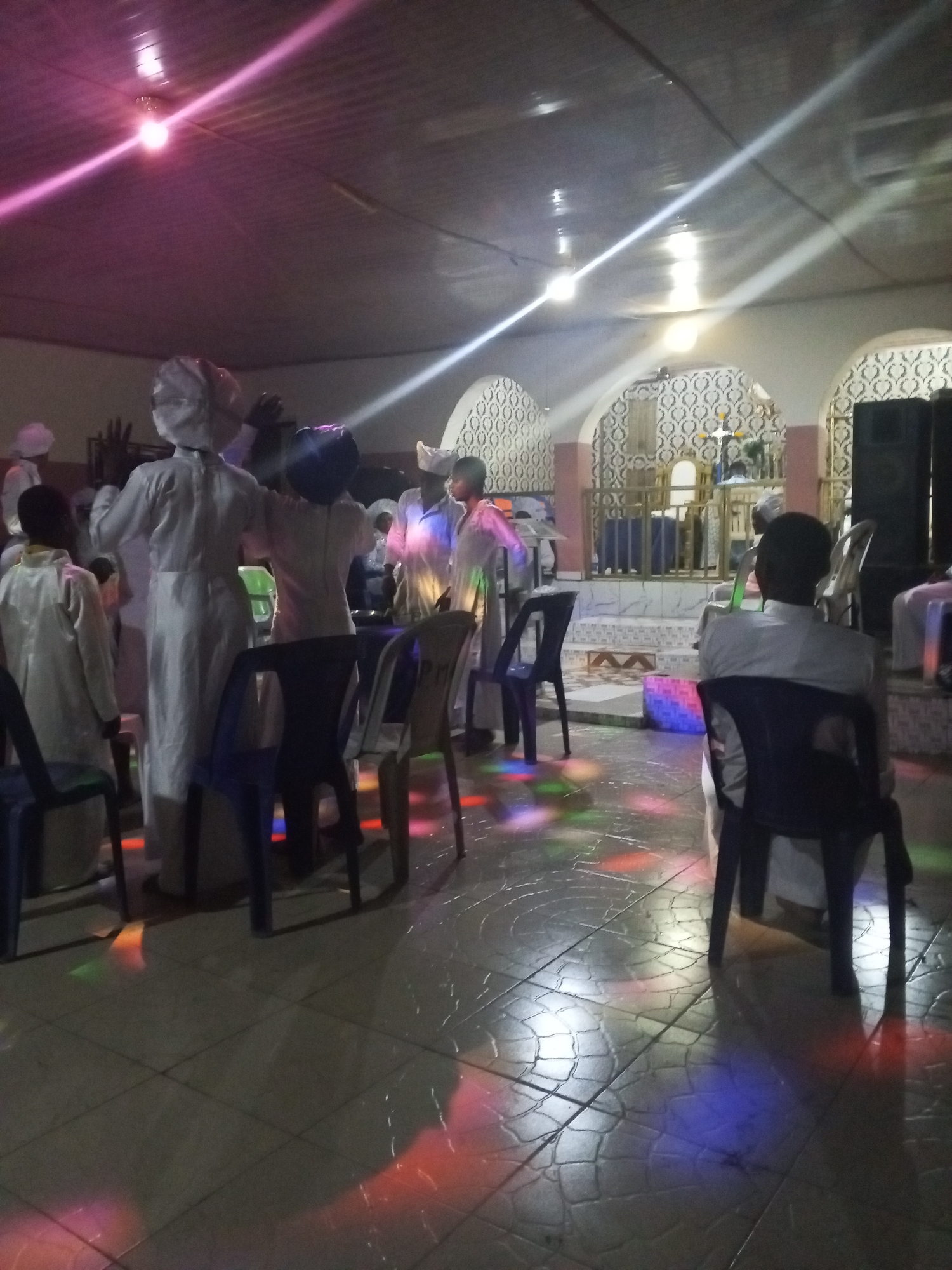 The image depicts a gathering inside what appears to be a church or religious hall in Lokoja, Nigeria. Several people dressed in white robes and head coverings are standing and appear to be participating in a service or ceremony. Some individuals are seated on chairs. The background features a raised platform with decorative arches and religious iconography. The lighting includes fluorescent ceiling lights and colored stage lighting that casts patterns on the floor. The setting is somewhat informal, with chairs scattered around the room. Speakers are positioned on the platform.