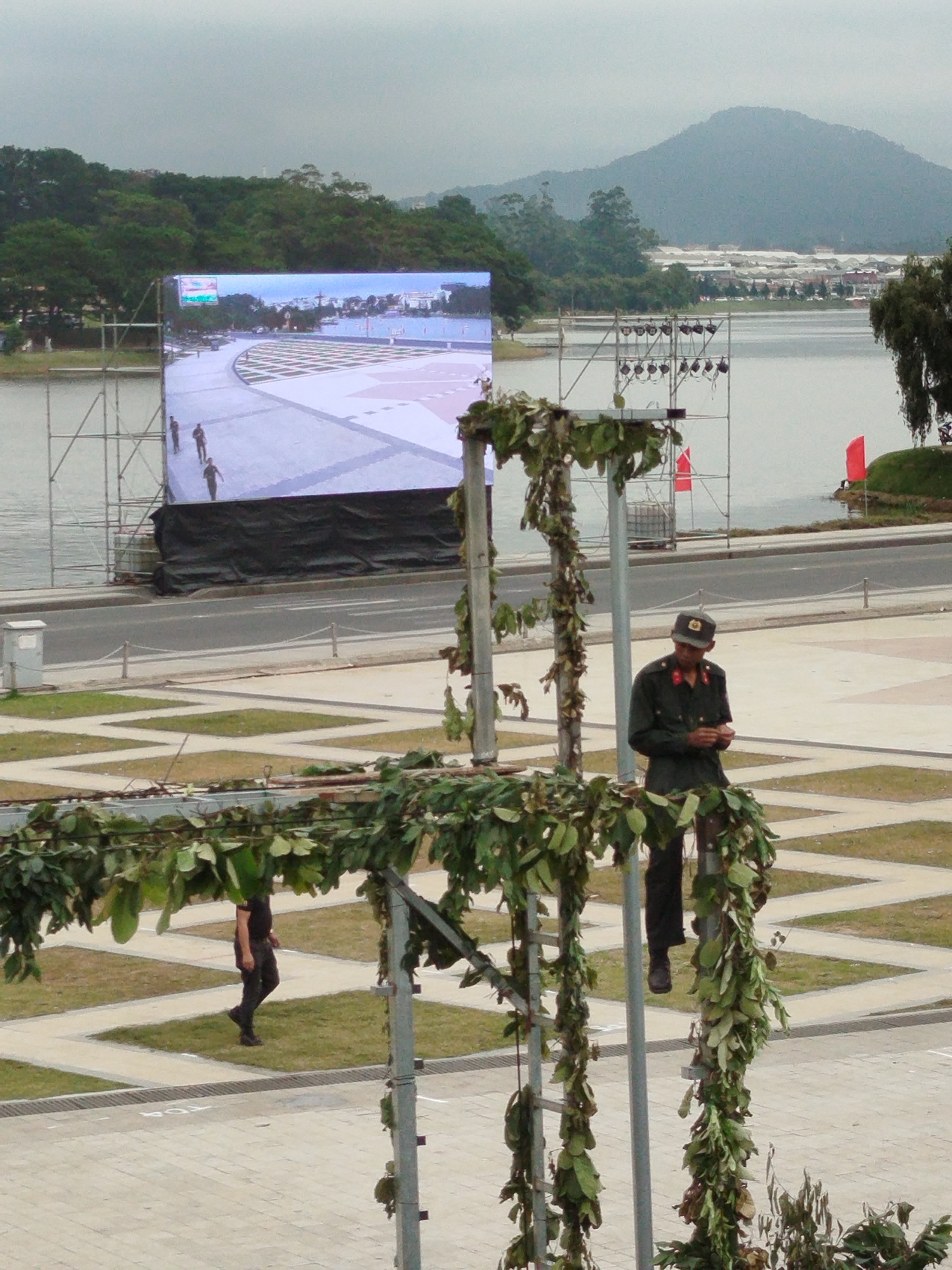 In Da Lat, Vietnam, a man in a military uniform stands near a large outdoor LED screen. The screen displays a landscape view, possibly of a city or park. The man, wearing a cap and a dark uniform with red accents on the collar and epaulets, appears to be looking at his phone. He is positioned on a metal structure adorned with greenery, suggesting a decorative or functional element of an event. To the left, another person in dark clothing walks away, their back to the camera. In the background, a body of water and a tree-covered hill are visible. Red flags are also present, attached to scaffolding near the water's edge. The overall scene suggests an outdoor gathering or event in a scenic location.