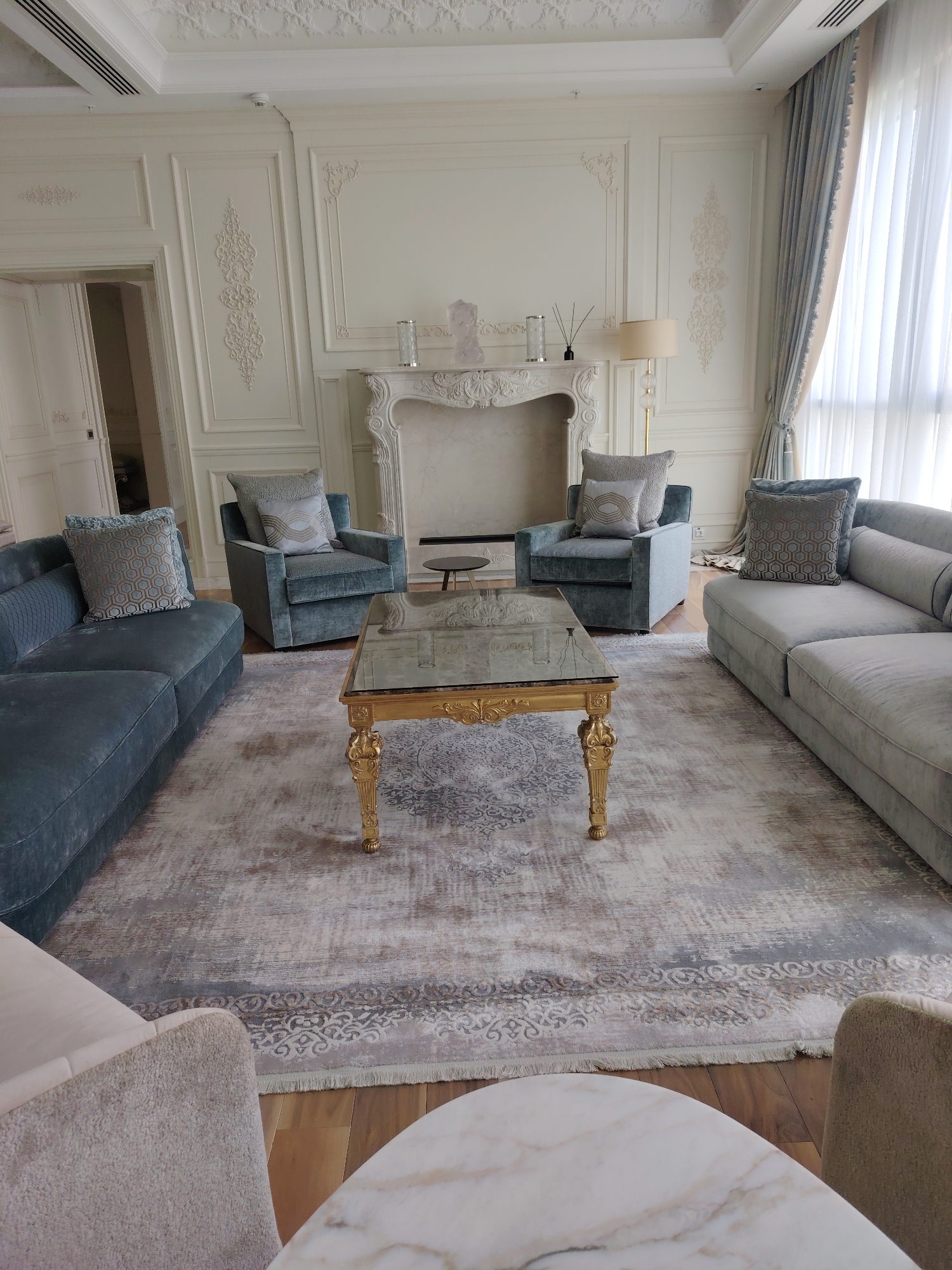 The image depicts a luxurious living room in Konigil, Uzbekistan, featuring ornate white wall molding and a grand fireplace. A plush, teal velvet sectional sofa is positioned on the left, complemented by a matching armchair to its right. Decorative pillows with geometric patterns in grey and teal adorn the seating. In the center of the room, a rectangular glass coffee table with a gold ornate base rests on a large, intricately patterned rug in muted grey and beige tones. Opposite the sectional, another teal velvet armchair is placed near a large window draped with light blue and beige curtains. A floor lamp with a cream shade stands beside the armchair. The room is brightly lit by natural light streaming from the window, and the overall ambiance is one of sophisticated comfort.