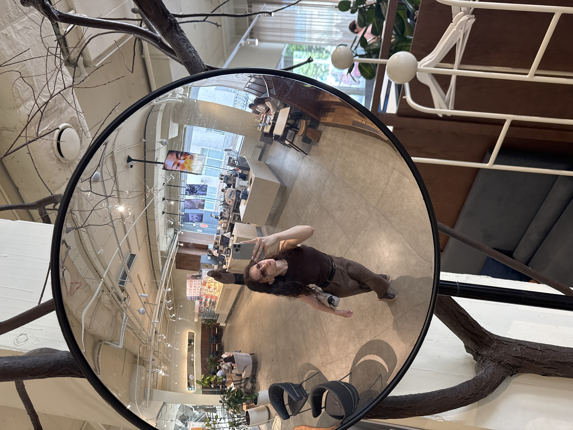 A woman is taking a selfie in a large, round mirror. She is wearing a dark brown tank top, brown pants, and sunglasses. She is holding a smartphone in her right hand and has a light-colored purse slung over her left shoulder. The mirror reflects a coffee shop interior. In the reflection, there are tables with people sitting at them, a counter with pastries and coffee machines, and plants. The woman is standing in the foreground of the mirror's reflection. The environment outside the mirror shows a wall with a large, dark, tree-like branch structure attached, and a white wall. This scene is located in the city of Yekaterinburg, Russia.