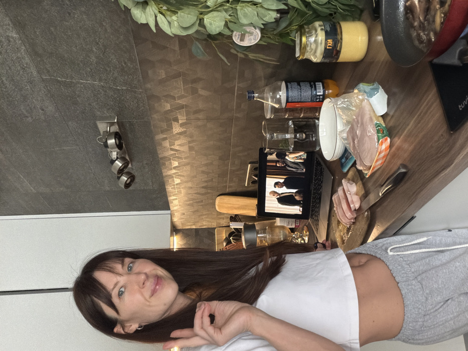 The scene depicts a contemporary kitchen in a residence located in Perm, Russia. A female subject with long dark brown hair and bangs, dressed in a white cropped t-shirt and grey sweatpants, stands on the left side of the frame, smiling directly at the camera. Her right hand is raised, forming an "OK" gesture with her thumb and forefinger.
The kitchen features a dark brown wood-grain countertop. Above it, a backsplash of brownish-gold tiles displays a geometric triangular pattern, illuminated by under-cabinet strip lighting. The upper wall consists of dark grey textured tiles, hosting a fixture with three silver cylindrical spotlights. White upper cabinets are partially visible on the far left.
Arranged on the countertop are multiple items. A silver laptop is open, displaying a video featuring four men in formal attire. To the laptop's left, a wooden cutting board holds sliced raw chicken breast, with a kitchen knife resting beside it. A package of raw chicken breast and a white ceramic bowl are nearby. A clear glass pitcher filled with water, a plastic bottle of orange-colored liquid, and a glass jar labeled "ГХИ" (Ghee) are also present. A glass with a stem, containing liquid and a dark object, stands near the laptop. In the foreground on the right, a red frying pan on a black induction hob contains food, possibly mushrooms, actively cooking. An artificial green leafy plant with a small white circular object attached is positioned on the far right. The scene captures the subject engaged in meal preparation while simultaneously viewing content on a laptop.