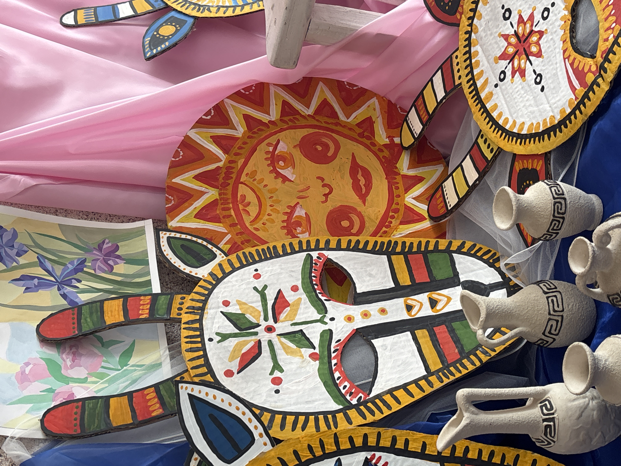 The scene depicts an indoor display of folk art elements, located in Tayshet, Russia. The primary subjects include multiple brightly painted, stylized cutouts and a collection of ceramic pottery.
In the central foreground, a large, white, mask-like cutout with black-outlined eye holes, a small mouth slit, and geometric patterns in red, green, yellow, and black is prominently displayed. It is adorned with two upward-curving, striped ear-like appendages. To its immediate right, a circular cutout portrays a stylized yellow sun with a human-like face rendered in red and orange tones, surrounded by radiating sunbeams. Partial views of additional painted cutouts are visible to the left and lower right, including one circular piece featuring a red and yellow starburst design, along with striped appendages similar to the main mask. These cutouts appear to be made from a rigid material, possibly cardboard.
Arranged on a blue fabric in the lower foreground are approximately six unglazed, light-colored ceramic vessels of varying sizes. These include pitchers with handles and smaller open-mouthed vases, many adorned with a dark, maze-like or geometric pattern around their midsections.
The background consists of a blue fabric drape on the left, two distinct watercolor paintings (one depicting pink roses, the other purple irises) centrally placed, and a draped pink satin fabric on the right. A partial view of a light-colored, possibly wooden, stool or small table is visible under the pink fabric on the far right. A translucent white netting or sheer fabric is draped throughout the display, particularly over parts of the background and foreground. The items are arranged in a static configuration, indicative of an exhibition or decorative setup.