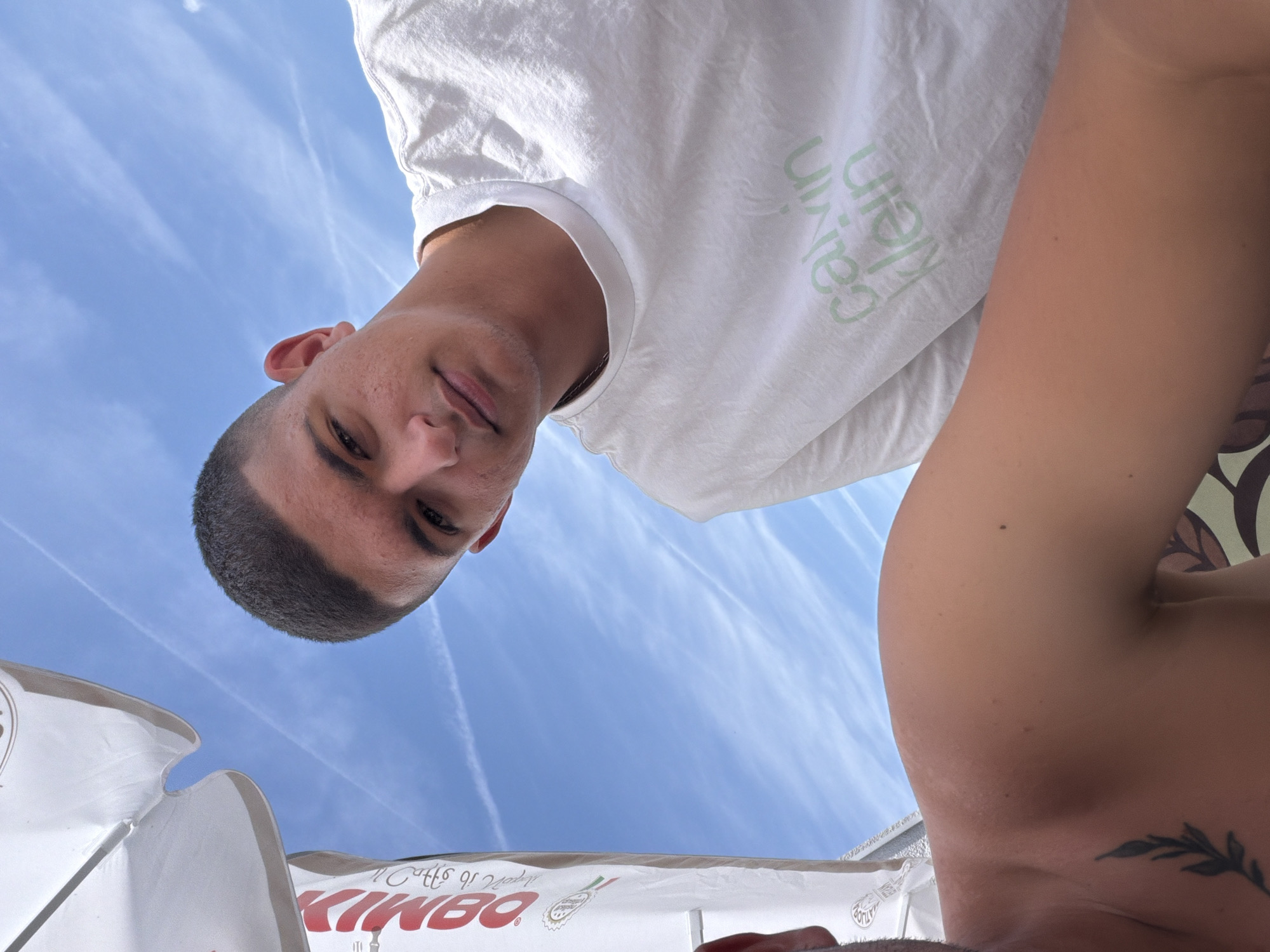 The image is a low-angle shot, looking upwards, and appears to be rotated approximately 90 degrees counter-clockwise. The main subject is a young man, positioned in the upper left portion of the frame (when viewed rotated), looking directly down at the camera with a neutral expression. He has short, dark, closely-cropped hair and dark eyes. He is wearing a plain white short-sleeved t-shirt with the brand name "Calvin Klein" printed in light green letters on the chest, appearing upside down and reversed due to the image orientation.
The background is a vibrant, clear blue sky with several distinct, thin white contrails from airplanes, indicating a bright, sunny day. To the right of the primary subject, a bare shoulder or upper arm of another individual is partially visible, featuring a dark-ink tattoo of a leafy branch.
In the bottom left of the frame, a white and red umbrella or canopy-like structure is present. On this structure, the word "KIMBO" is prominently displayed in large, red, block letters, also appearing reversed and upside down. Below "KIMBO," smaller text is visible, which seems to read "il caffè di Napoli."
The overall setting is outdoors on a clear, sunny day, suggesting a leisure environment such as a beach, poolside, or outdoor cafe, given the presence of the umbrella and open sky.