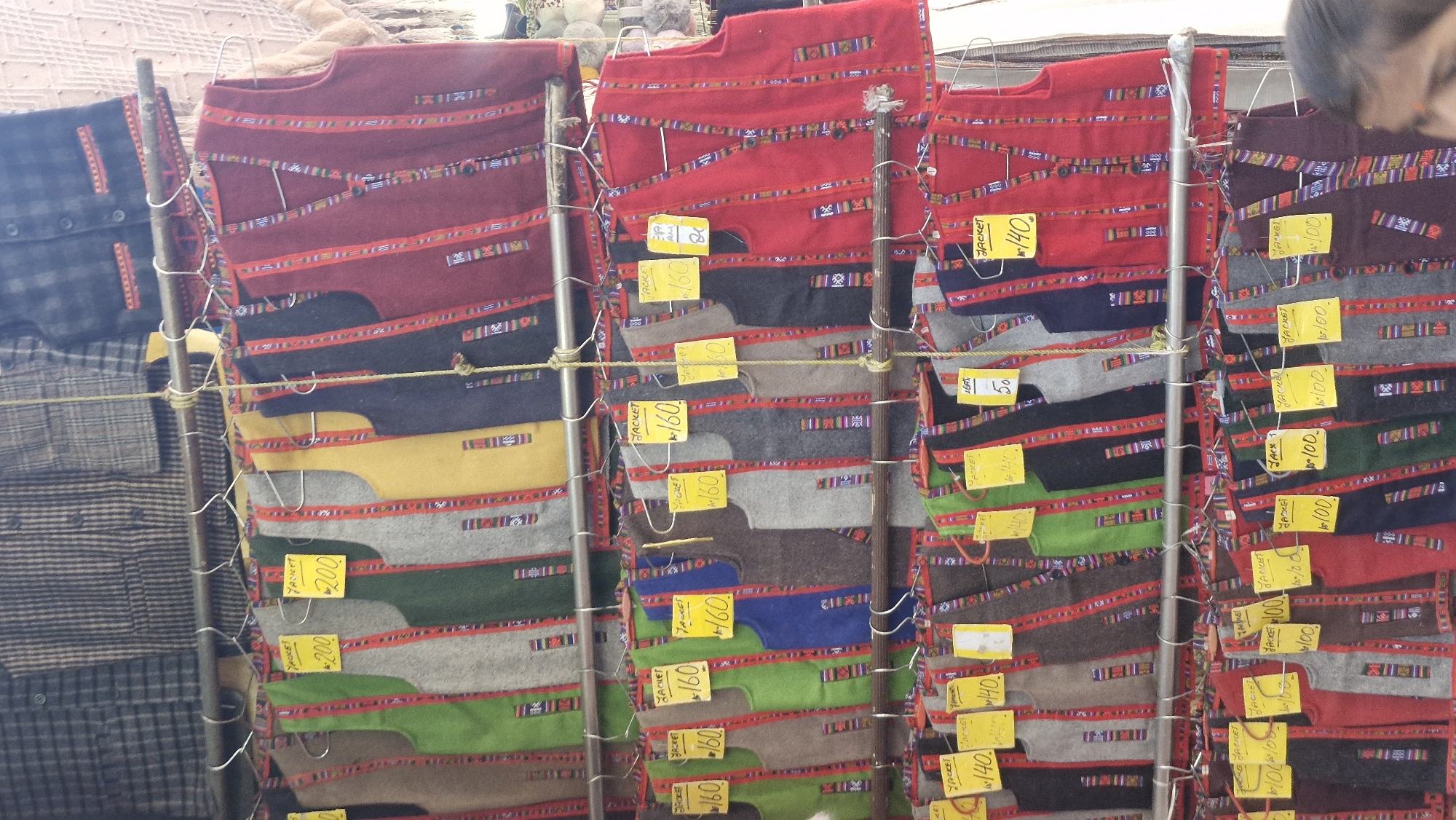 The image shows a stall displaying numerous colorful, sleeveless vests, likely for sale. The vests are arranged in rows on hangers, with some bearing yellow price tags. The background suggests an outdoor market or shop, possibly in Vashisht, India, given the context provided. The vests are predominantly in solid colors such as red, green, yellow, grey, and dark blue, and feature intricate woven patterns along the edges and down the front. Some vests have buttons. In the foreground, the shoulder and hair of a person are visible, indicating a shopper or vendor is present. The lighting appears to be natural daylight.