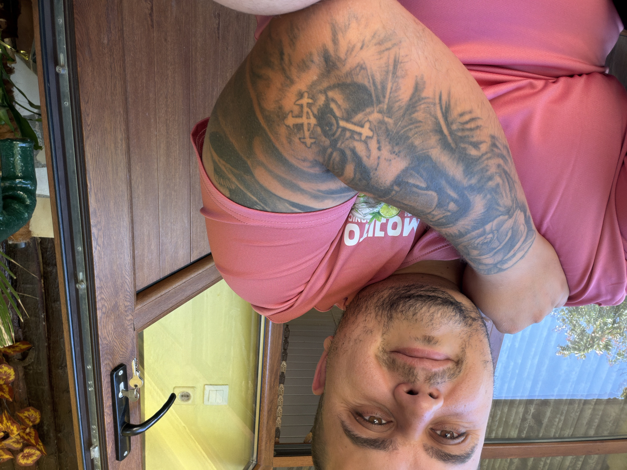 An individual is posing for a selfie, possibly in Kaolinovo, Bulgaria, seated outdoors near a wooden door frame. The individual is wearing a pink t-shirt with white text that appears to be "MOJITO" and features a graphic of limes and mint. They have extensive tattoos on their left arm, including what looks like a lion's face and a cross symbol. The background shows a wooden door with a black handle and keys, a yellow wall with light switches, and some greenery. The lighting suggests it is daytime.