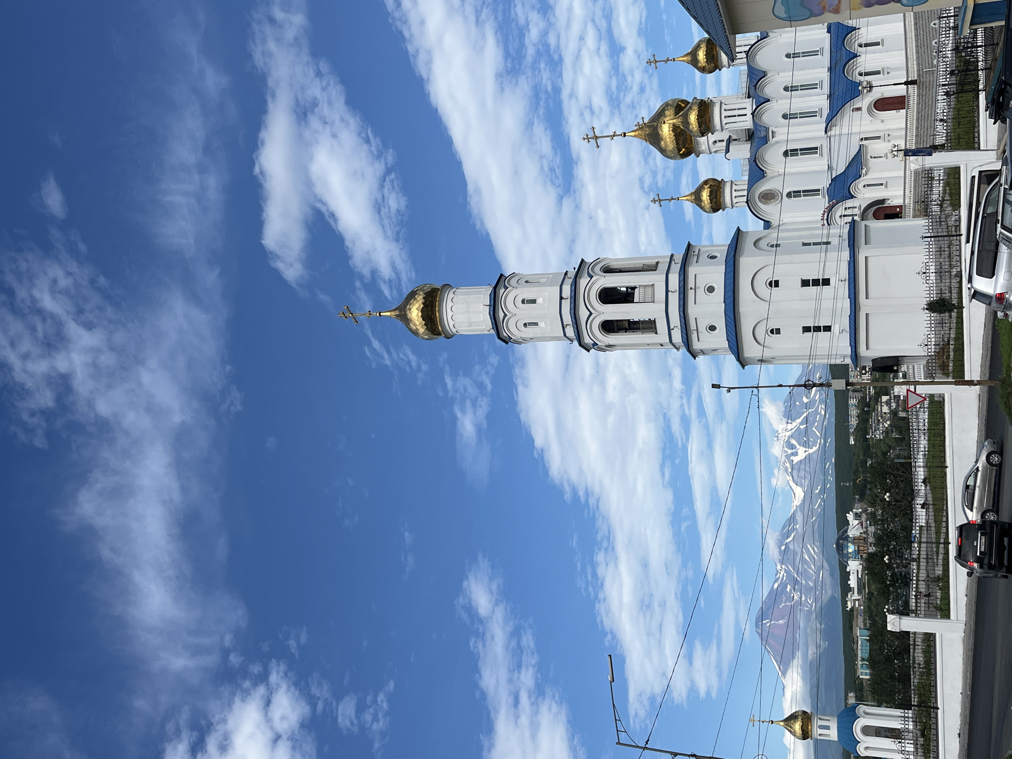 In Petropavlovsk-Kamchatsky, Russia, a bright, sunny day unfolds with a clear blue sky dotted with fluffy white clouds. Dominating the foreground is a magnificent white Orthodox church with a tall, slender bell tower featuring a golden dome topped with a cross. Adjacent to it, the main church structure boasts multiple golden domes, also adorned with crosses, and striking blue rooflines. A smaller, detached bell tower with a blue roof and golden dome stands to the left, partially obscured by a street lamp.
In the background, a range of majestic, snow-capped mountains rises, their peaks partially shrouded by clouds. The city sprawls between the church and the mountains, with a mix of buildings, trees, and roads visible. A black SUV and a silver sedan are parked on the asphalt road in the lower part of the image, while other vehicles are visible further down the street. A white fence lines the perimeter of the church grounds. The overall scene captures a blend of religious architecture and the striking natural beauty of the surrounding volcanic landscape.