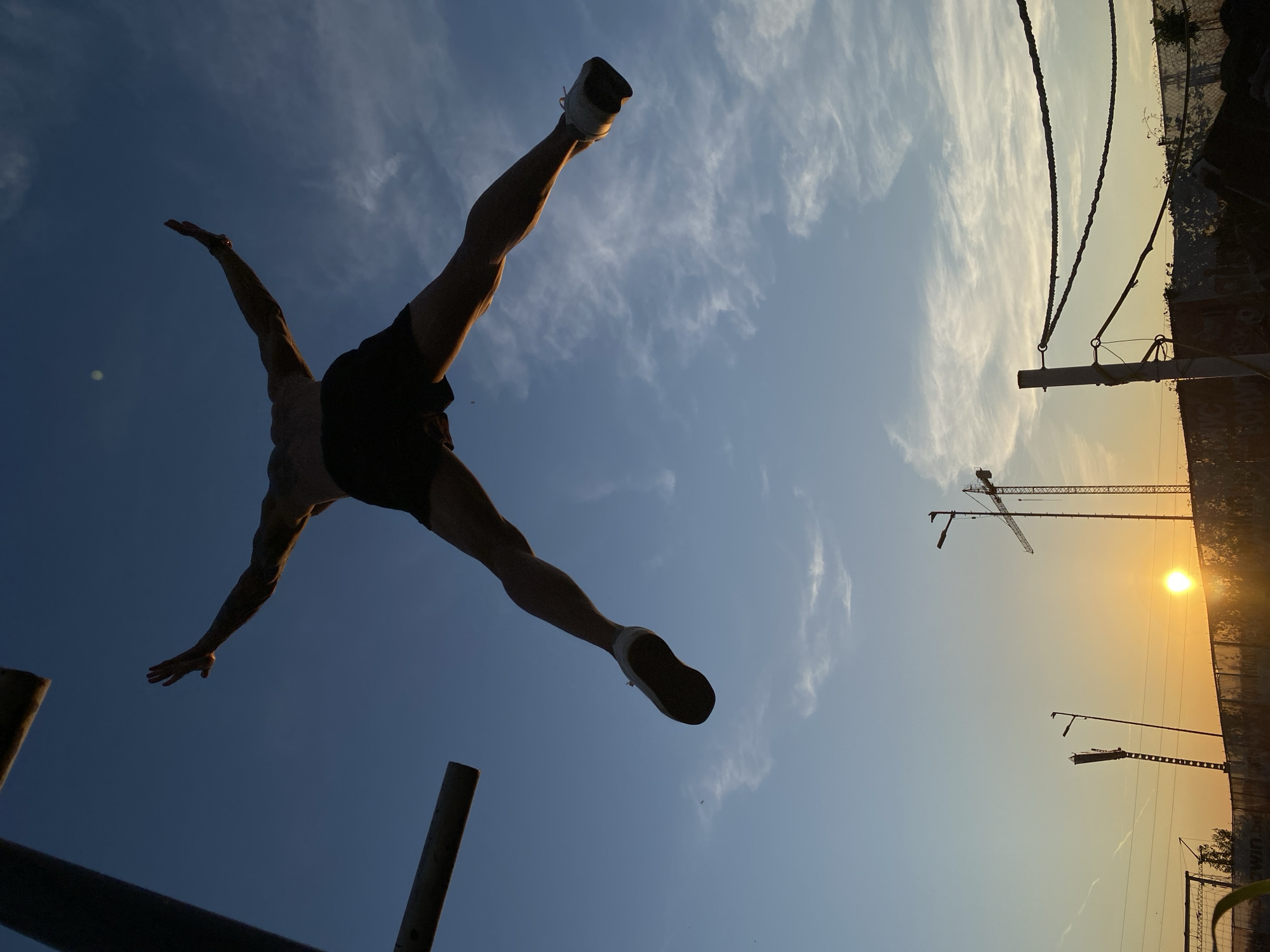 An adult male, silhouetted against a twilight sky, is captured mid-air in a star jump. He is wearing dark shorts and white athletic shoes. His arms are outstretched to the sides, and his legs are spread wide. The sky above is a gradient of deep blue to lighter hues, with wispy clouds scattered across it. In the background, a sunrise illuminates a construction site with cranes and scaffolding. The sun is visible on the horizon, casting a warm glow. The scene suggests an outdoor athletic activity or performance. The location is the city of Pleven, Bulgaria.