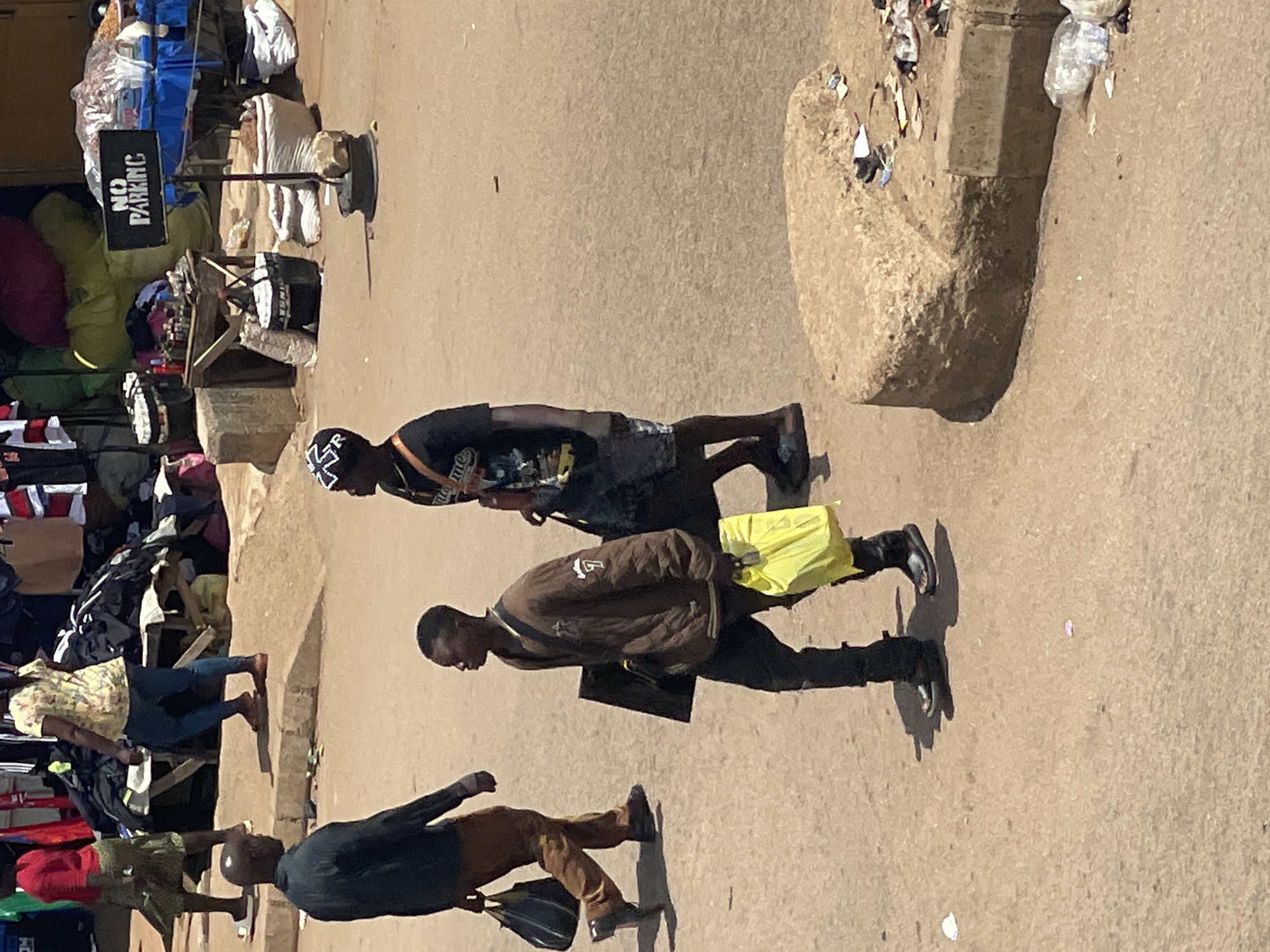 The image depicts an outdoor marketplace scene, likely in a city, with a focus on individuals walking on a dirt or gravel ground. In the foreground, two men are walking. The man on the left is wearing a black long-sleeved shirt, brown pants, and black shoes, carrying a black bag over his left shoulder. The man on the right is wearing a brown quilted jacket over a t-shirt, dark pants, and dark sandals. He is carrying a flat black object under his left arm and a bright yellow plastic bag in his right hand. To the left of these two men, a third man in a black shirt and brown pants is walking away from the camera. In the background, several other people are visible, including a woman in a yellow floral shirt and blue pants, and another woman in a red top and green skirt. The marketplace is filled with stalls and piles of goods, including clothing and bags. A "NO PARKING" sign is prominently displayed on a stand. The environment is dusty and appears to be a busy, informal commercial area.