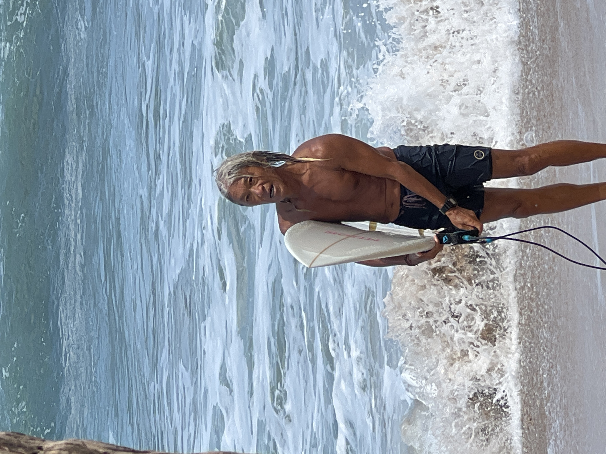 An adult male surfer with long, graying hair stands on the wet sand of a beach, holding a surfboard. He is wearing black swim trunks and a watch on his left wrist. The ocean behind him is choppy with white water breaking near the shore. The image is taken from a medium distance, showing the surfer from the knees up. The background consists of the ocean and a rough, sandy shoreline. The lighting suggests it is daytime.