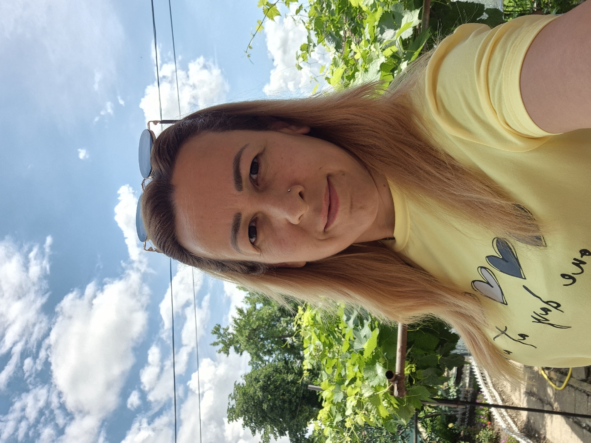 A medium-shot, selfie-style photo shows a woman with long, blonde hair and a yellow t-shirt with a heart design on it. She is standing outdoors in front of a background of lush green foliage and a bright blue sky with scattered clouds. Sunglasses are perched on top of her head. The location is not specified as being in a city or any particular place.