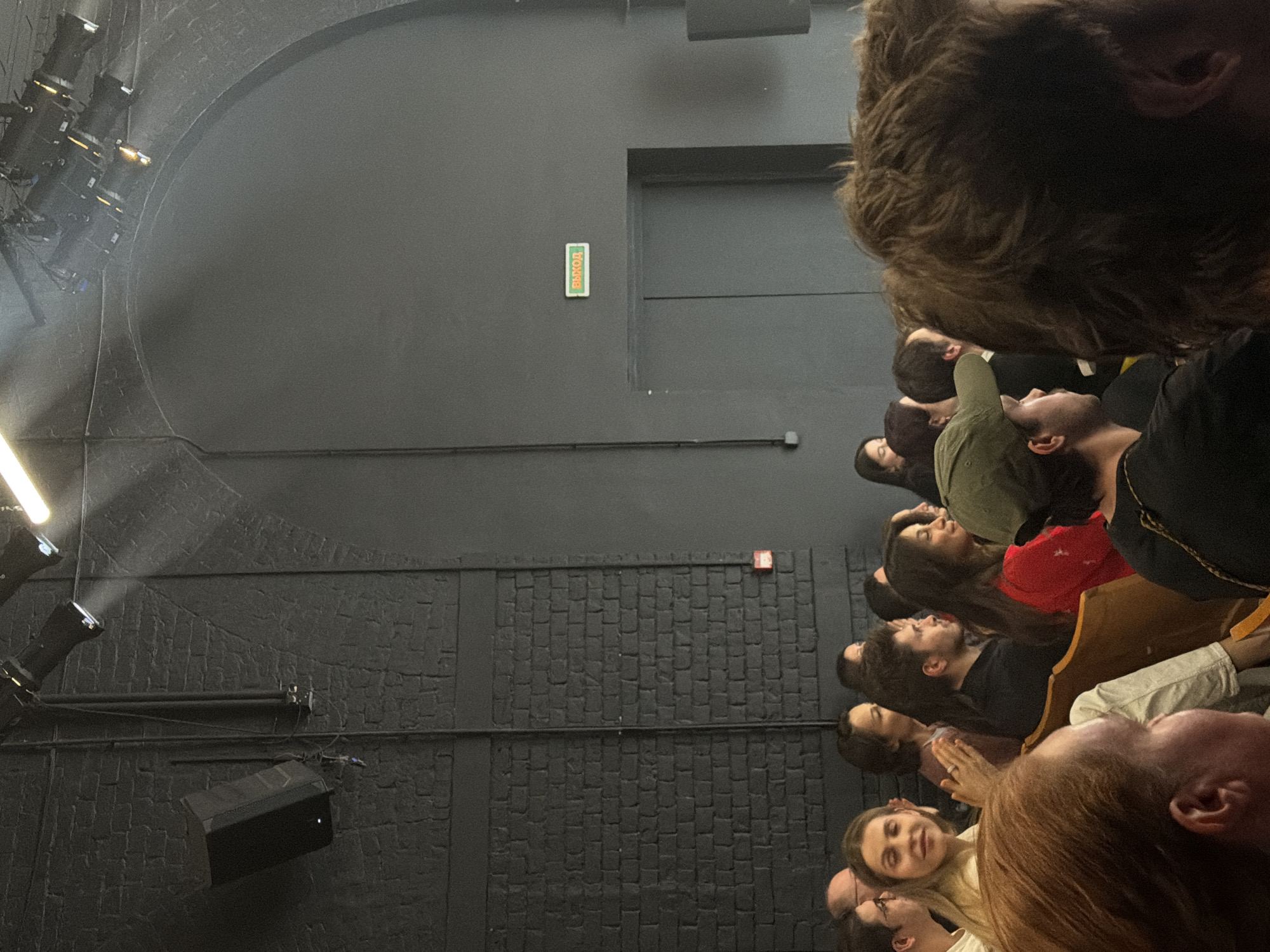 This image captures an indoor scene, likely a theater or performance venue, from the perspective of an audience member looking towards the stage. The photo appears to be taken slightly from above and to the right of the main group of spectators.
The primary subjects are people forming an audience, seated in rows and predominantly looking forward and slightly upwards towards an unseen focal point or stage to the left of the frame. In the immediate foreground, the back of a person's head with thick, dark brown hair is prominent, along with a glimpse of another person with reddish-brown hair. Further back, faces of numerous individuals of varying ages and appearances are visible, many exhibiting expressions of attentiveness, interest, or slight smiles.
The setting is characterized by dark grey and black walls. Some sections of the wall feature a smooth finish, while others have a distinct brick-like texture. On the left side of the frame, several stage lights are mounted on rigging, casting beams of light. A black speaker box is also mounted on the wall. Towards the rear of the venue, a green and white sign with the Cyrillic text "ВЫХОД" is clearly visible, indicating an exit. A small red box, possibly a fire alarm or sensor, is also present on a textured wall section.
The activity taking place is an audience watching a performance, lecture, or presentation. The controlled lighting and stage equipment suggest a professional artistic or educational event. Given the "ВЫХОД" sign and the provided context, the location is Moscow, Russia. The atmosphere appears focused and engaging.