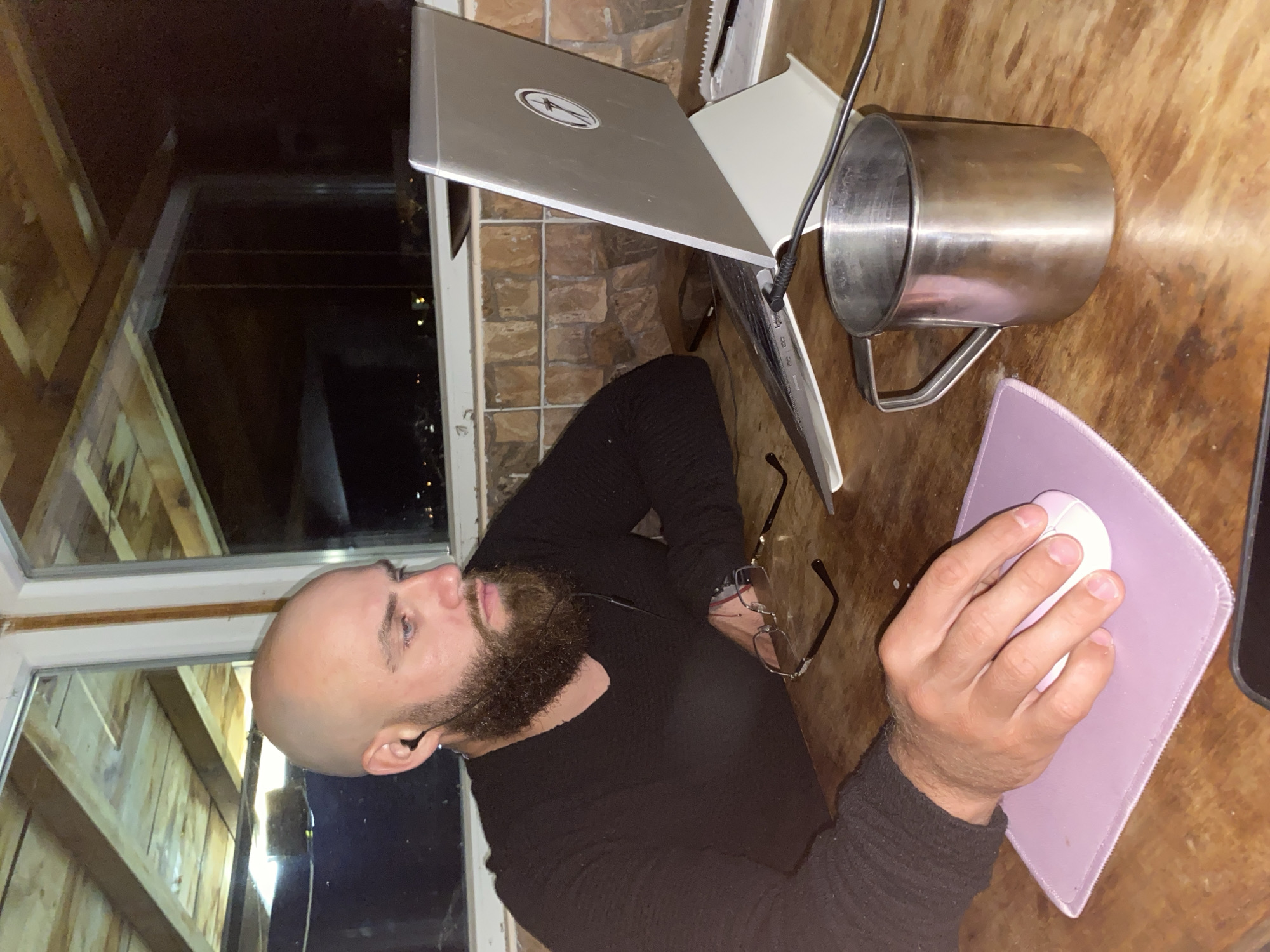 An indoor eye-level shot, rotated 90 degrees clockwise, shows a man seated at a wooden table, engaged in a computer-related activity at night. The man, with a bald head and a full, dark beard, wears a black long-sleeved shirt. He has an earbud in his left ear and appears to be looking forward or slightly upward. His right hand rests on a white computer mouse, positioned on a light purple mouse pad.
On the table in front of him, a silver laptop stands open on a light-colored riser. A circular sticker, resembling a stylized globe or star, is visible on the laptop's lid. To the right of the laptop is a large, polished metal mug with a handle. Black cables are seen around the laptop and mug. A pair of black-framed eyeglasses rests on the table near the man's left arm.
The background features a large window to the man's left, looking out onto a dark scene with faint distant lights, indicating it is nighttime. The room has warm-toned wooden paneling on the walls and possibly the ceiling, with a section of brick-like wall visible behind the laptop. The overall setting suggests a cozy, possibly rustic, indoor workspace in Bankya, Bulgaria. No explicit text is clearly visible.