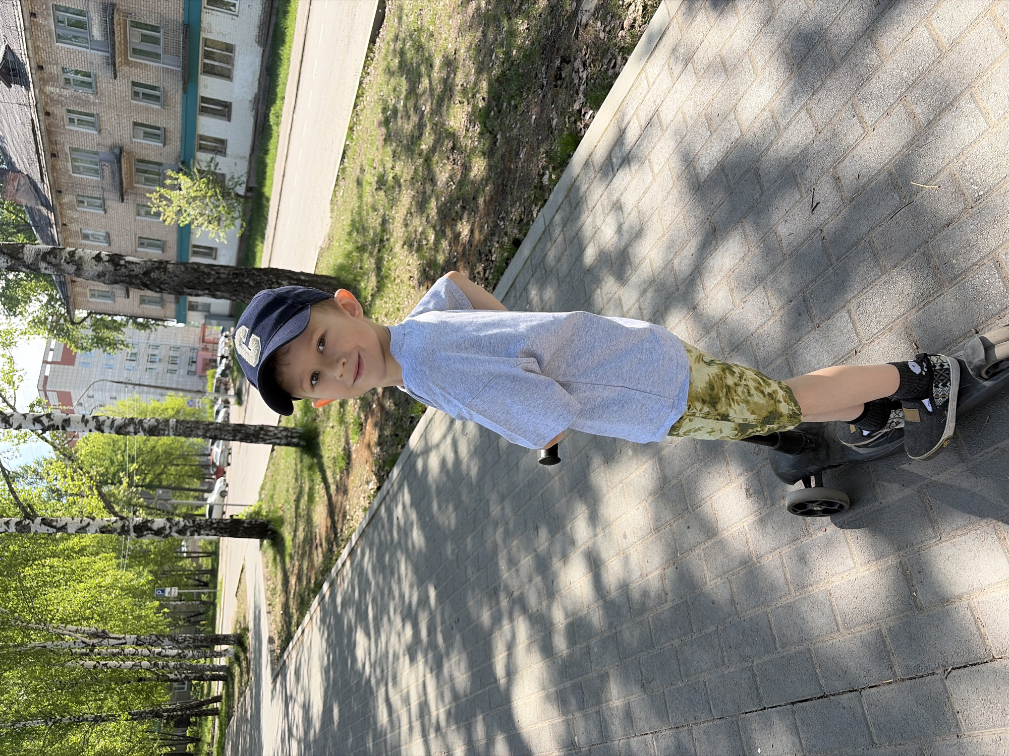 A young boy wearing a blue baseball cap, a grey t-shirt, and patterned shorts is riding a scooter on a paved walkway. The walkway appears to be made of brick or stone, and there are shadows cast upon it, likely from trees. The boy is looking towards the camera. To the left of the walkway are trees, and beyond the trees, there are buildings. The scene is in the city of Chaikovsky, Russia.