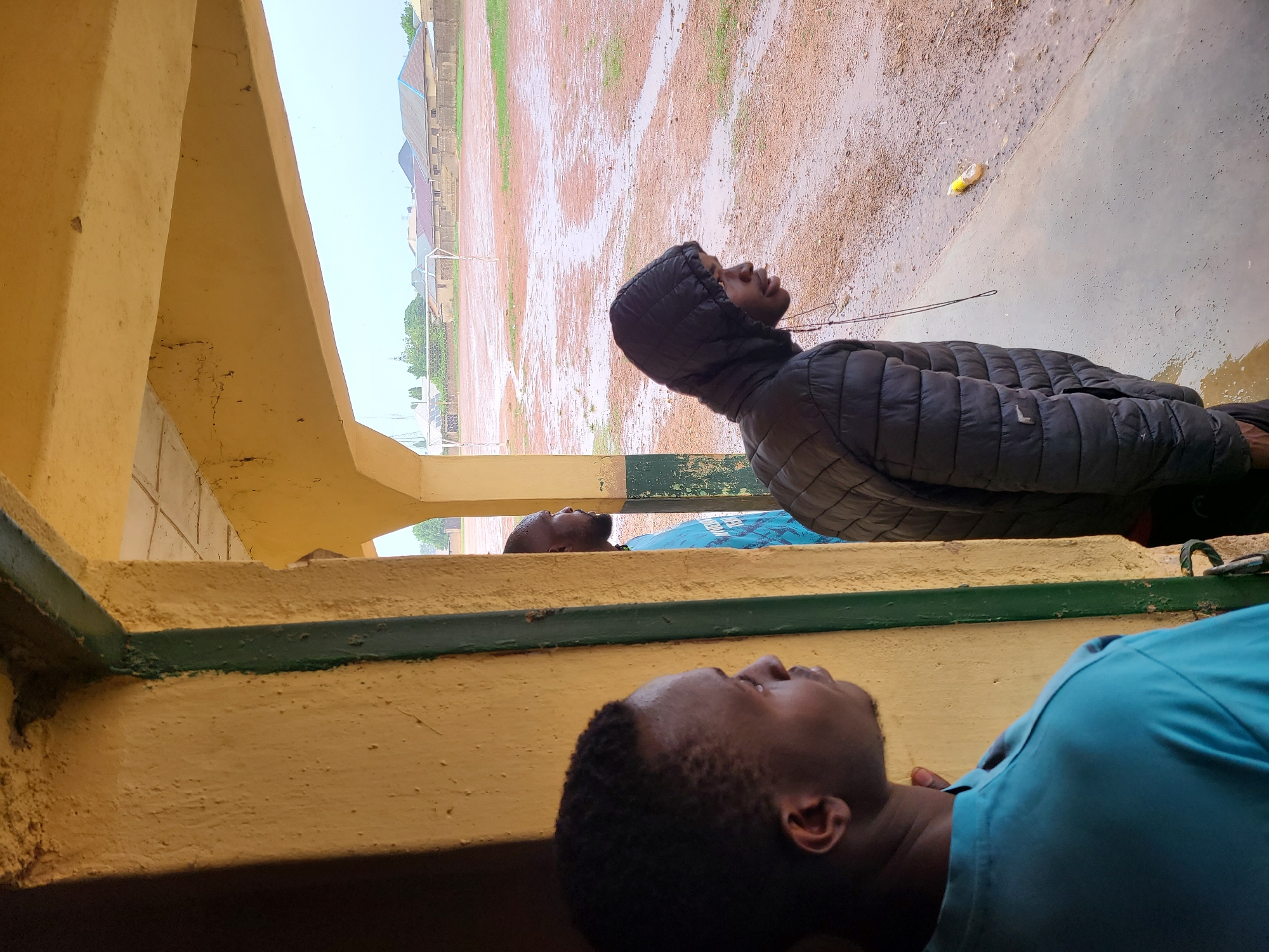 The image captures an outdoor scene featuring three young men and a large, muddy field, presumably after rainfall. The perspective is from beneath a yellow-painted concrete structure, possibly a balcony or overhang, with a dark green trim line.
In the foreground, a man with short, dark hair, wearing a light blue short-sleeved top, is partially visible on the right, looking upwards and slightly away from the camera. In the mid-ground, a second man stands further back, wearing a black hooded, padded jacket. He also has a dark complexion and is looking upwards towards the distant field, with a black earphone cord dangling from his ear. A third man is partially visible in the background, between the foreground and mid-ground figures, wearing a blue top and also looking upwards.
The ground beyond the structure is a wide expanse of red-brown earth, heavily saturated with water, forming numerous puddles and mud patches. In the distance, on the left side of the field, a white soccer goalpost is visible. Behind the field, various buildings with different roof styles can be seen under an overcast sky. A yellow plastic bottle or piece of debris lies on the muddy ground near the man in the black jacket.
The scene suggests a moment of contemplation or observation following a period of rain, given the wet and muddy conditions of the field. The men appear to be looking out at the environment or up towards the sky. No visible text is present in the image.