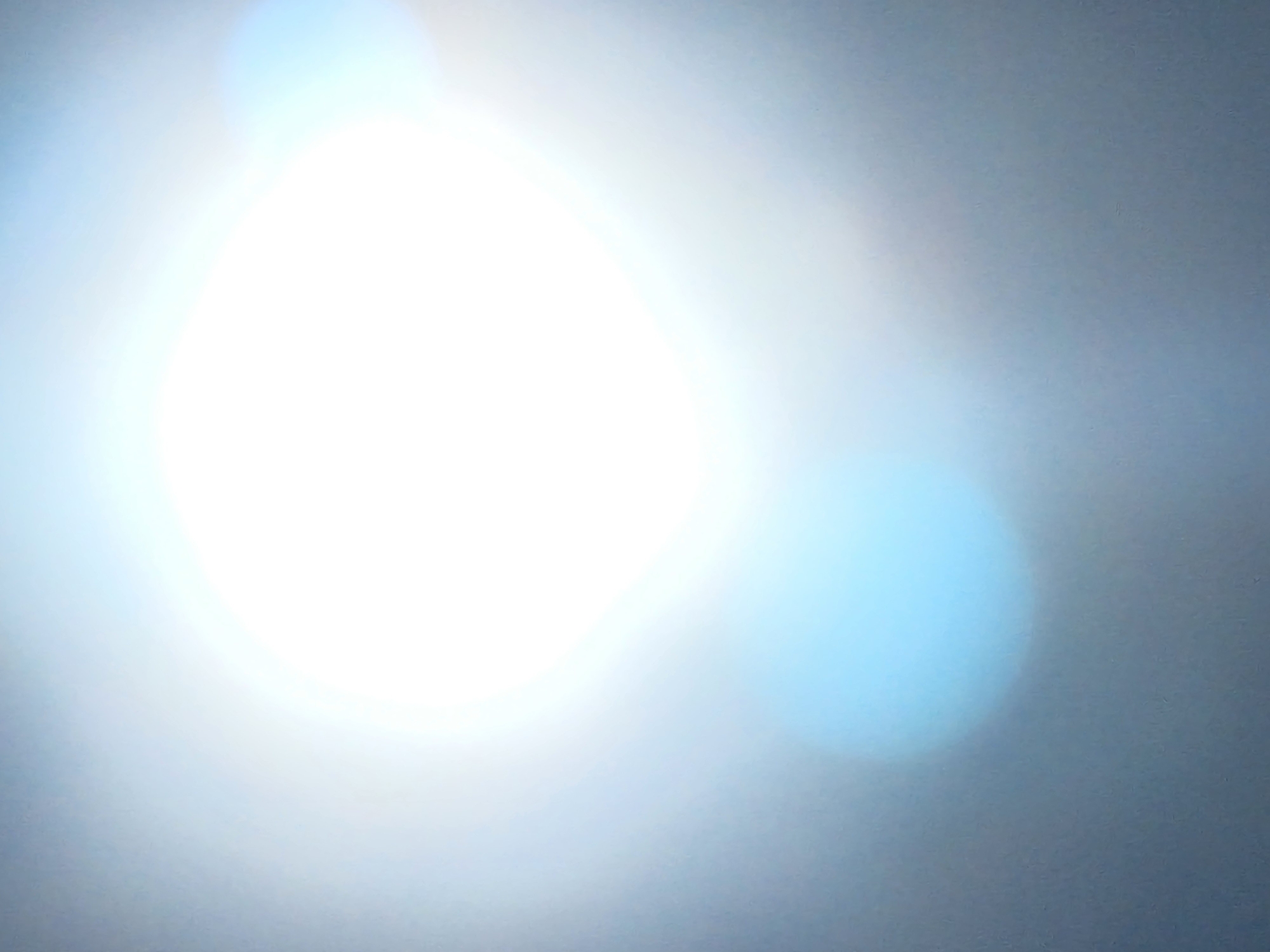 The image is heavily overexposed and blurred. The primary feature is a large, bright white blob, likely a light source or reflection. There is a softer, light blue orb to the right and below the bright area. The background appears to be a muted gray or blue-gray color. No specific location, people, subjects, objects, actions, or interactions can be identified due to the poor quality and overexposure of the image.