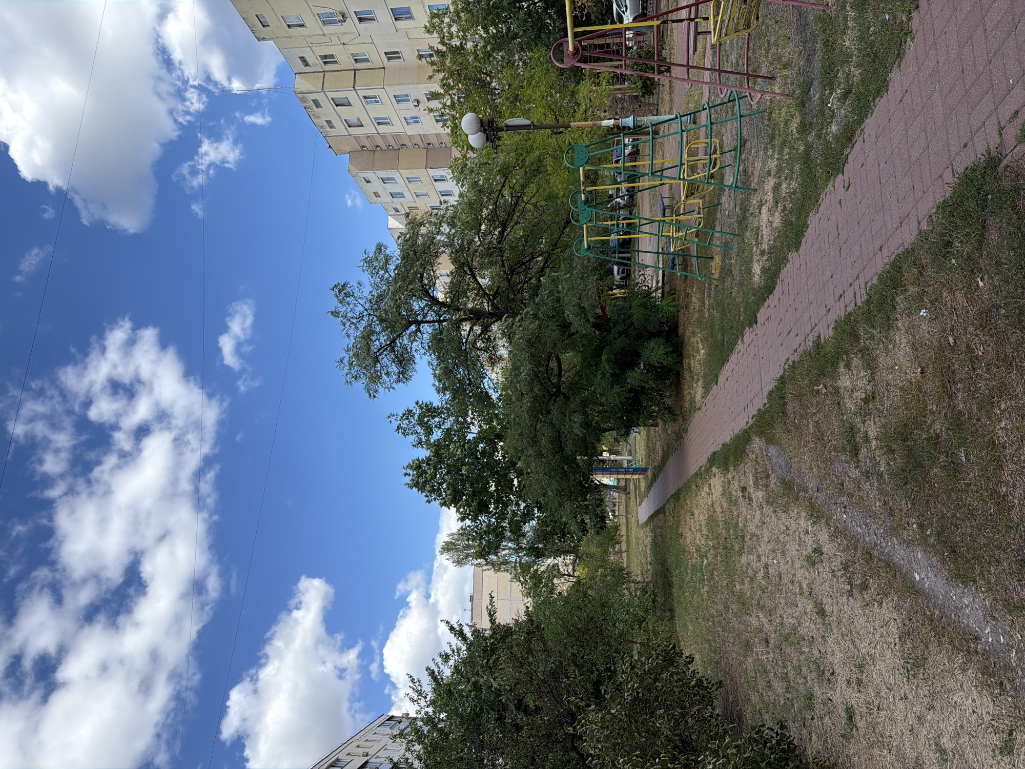 The image shows a courtyard in Kyiv, Ukraine, on a sunny day with scattered clouds. A paved walkway, made of red brick pavers, leads through a grassy area with dry patches. On the right side of the walkway, there is a playground with several swing sets, painted in yellow and green, and some red metal frames. In the background, there are tall, multi-story apartment buildings, typical of Soviet-era architecture, with light yellow facades and numerous windows. Trees with green foliage are interspersed throughout the scene, some taller than the playground equipment and others smaller bushes. The sky is a bright blue, with fluffy white clouds. The overall impression is of a typical urban residential courtyard.