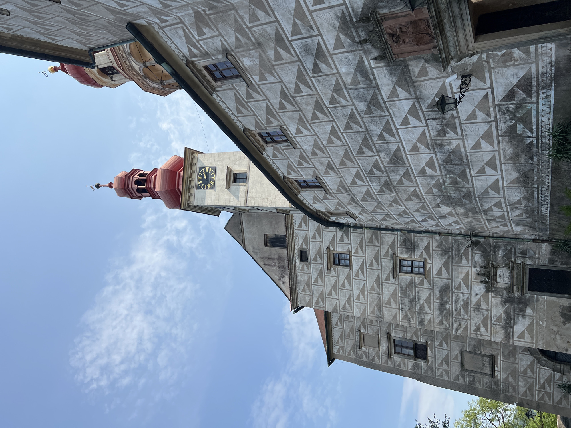 The image captures a low-angle view of the Náchod Castle in Náchod, Czechia, under a bright blue sky with scattered clouds. The dominant feature is the castle's architecture, characterized by a unique sgraffito technique on its exterior walls, creating a distinctive geometric pattern of light and dark grey triangles.
Several sections of the castle are visible. To the left, a tall, grey tower with a red, onion-shaped dome and a flagpole stands prominently. This tower features a clock face with Roman numerals. Adjacent to it, and slightly to the right, is another tower with a similar red dome, showcasing arched openings and decorative elements. The main body of the castle wall, to the right and center of the frame, is adorned with the aforementioned sgraffito pattern. This wall has several windows, some with dark frames, and an ornate stone relief above a doorway. A black, wrought-iron lantern is mounted on this wall.
The foreground shows the base of the castle, with a dark doorway and some greenery. The overall impression is one of historical grandeur and architectural uniqueness.
