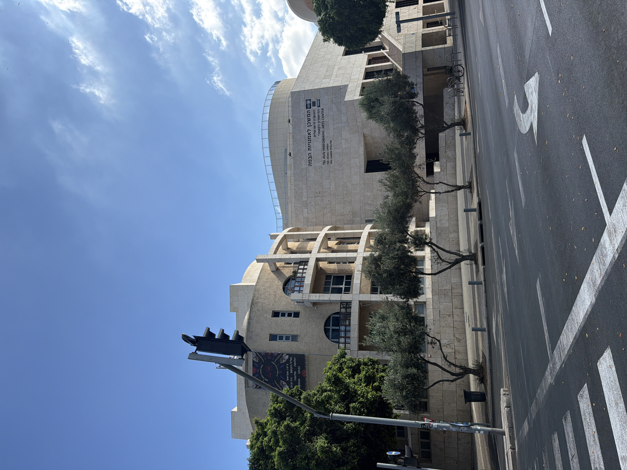 The image shows a street scene in Tel Aviv, Israel, featuring the Tel Aviv Performing Arts Center. The building, constructed with light-colored stone and curved architectural elements, is visible on the right side of the frame. Text on the building reads "TEL AVIV PERFORMING ARTS CENTER THE ISRAELI OPERA THE CAMERI THEATRE" and in Hebrew, "המשכן לאומנויות הבמה". A traffic light stands on the left, attached to a pole with a banner displaying "40". Several olive trees with bare branches line the sidewalk in front of the building. The street in the foreground has white crosswalk markings and a white arrow indicating a turn. A bicycle rack with a parked bicycle is visible near the building's entrance. The sky is blue with scattered white clouds.