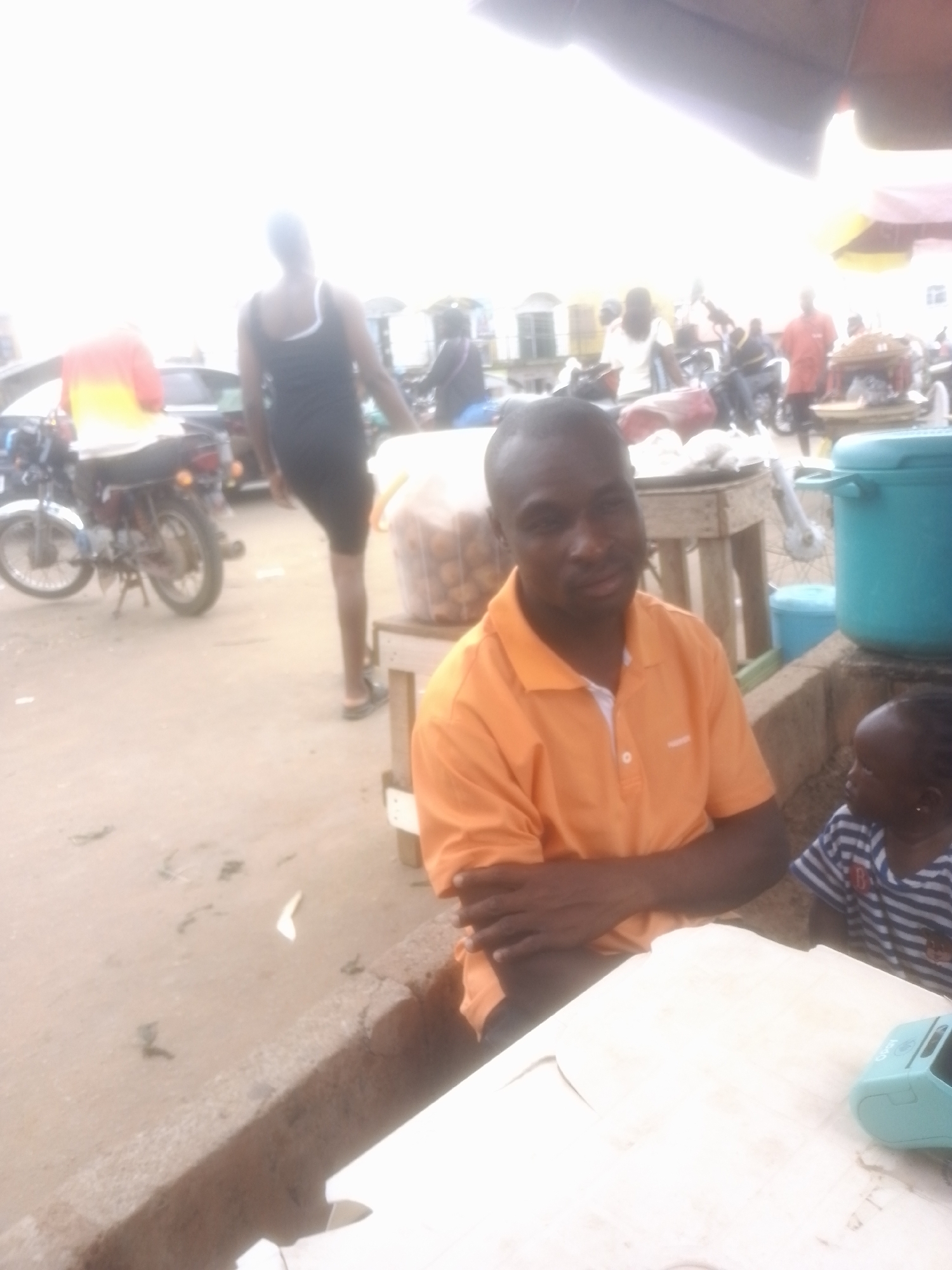 The image depicts a scene in Gwong, Nigeria, likely during daylight hours as indicated by the bright, washed-out sky, suggesting a very bright day, possibly with some haze. The setting appears to be an outdoor market or roadside area, characterized by dirt ground, concrete structures, and various objects associated with commerce and daily life.
In the foreground, a man wearing an orange polo shirt with a white collar is seated, looking towards the viewer. His arms are crossed, and he has a neutral expression. To his right, a young child wearing a striped t-shirt is also present, looking away from the camera. The child's facial features are partially visible. In front of them is a white table, upon which a small, light blue electronic device, possibly a point-of-sale terminal or a small speaker, is placed.
Behind the seated man, there are several other individuals and objects. A woman in a black dress stands with her back to the camera, facing away into the market. Further in the background, there are motorcycles, carts, and other people engaged in various activities, typical of a bustling marketplace. A large blue plastic container is visible to the right of the seated man. The overall atmosphere suggests a lively, everyday scene in a Nigerian community. The image has a slightly grainy quality and is overexposed in some areas, particularly the sky. No discernible text is visible in the image.