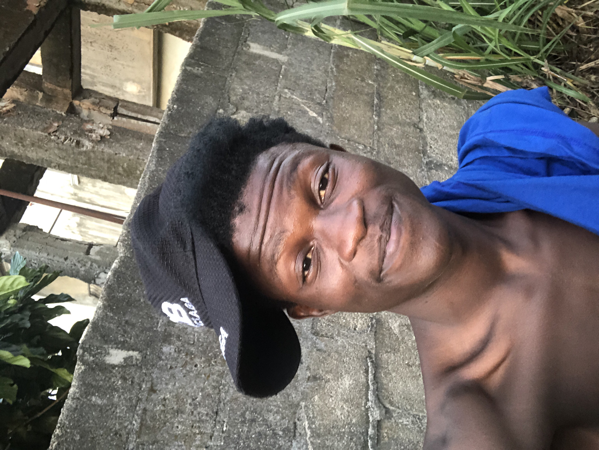 A close-up selfie of a young Black male is captured outdoors, with a grey concrete wall serving as the backdrop. The subject, wearing a black baseball cap with a white logo and a blue cloth draped over his shoulders, looks directly at the camera with a subtle smile. His dark, curly hair peeks out from under the cap. The lighting suggests it is daytime, possibly late afternoon, casting gentle shadows on his face. To the right of the subject, tall green plants are visible, adding a touch of nature to the urban setting. The overall impression is casual and informal, likely taken in a residential area or an alleyway in Ado, Nigeria.