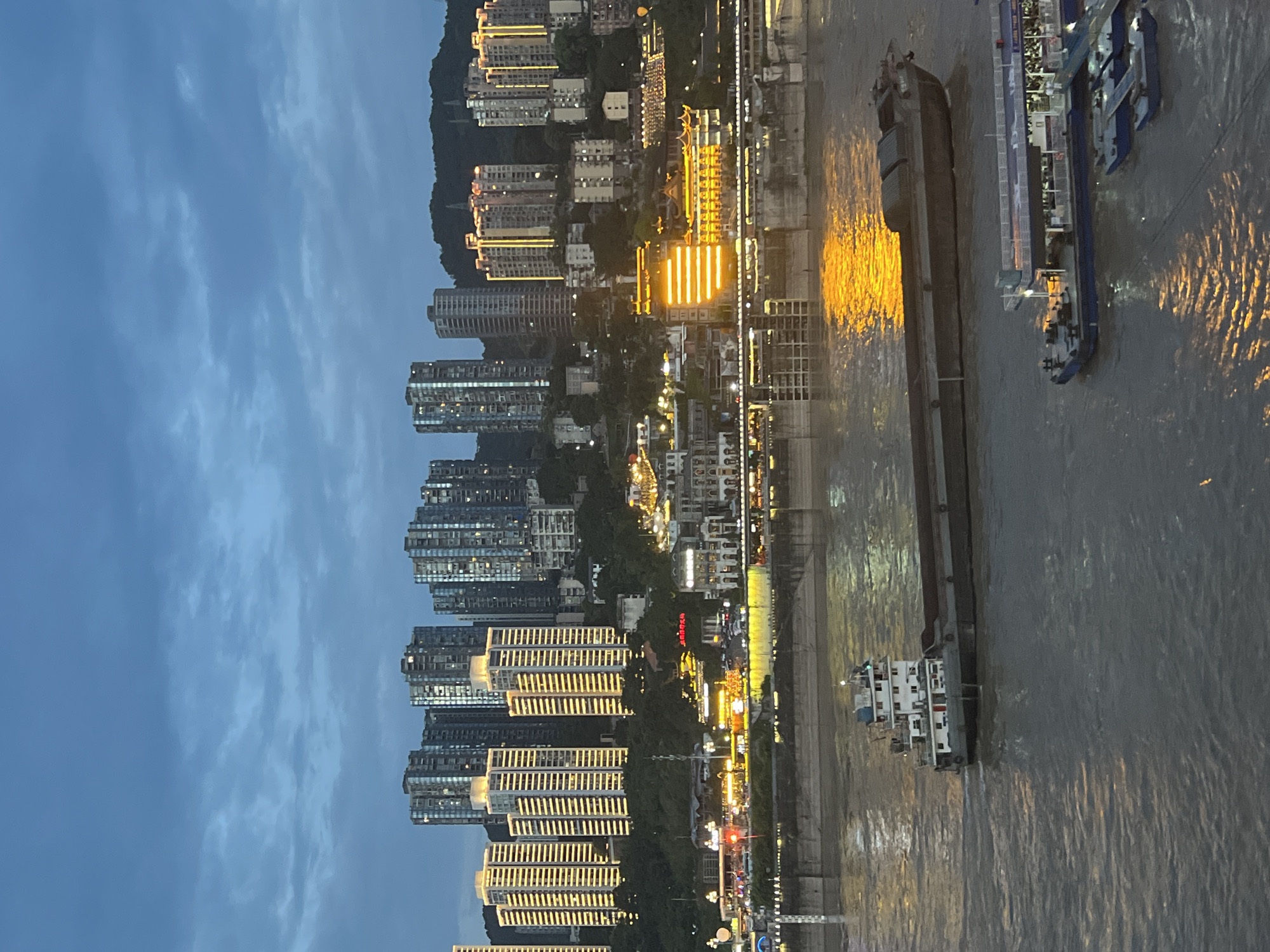 This is an aerial, eye-level shot of the Yuzhong District in Chongqing, China, at dusk. The sky is a deep blue with wispy clouds, illuminated by the city lights. The city's skyline is dominated by numerous high-rise buildings, some with brightly lit windows and others outlined with yellow lights. In the foreground, a wide, murky river flows, reflecting the golden lights from the city.
On the river, a long, flat barge is being towed by a tugboat. The tugboat, at the front of the barge, is white with a red trim on its cabin. Further down the river, two smaller boats are docked together at a pier. One of these boats appears to be a passenger ferry, with people visible on its deck. The buildings lining the riverfront are also illuminated, creating a vibrant contrast with the darkening sky. The scene depicts a bustling urban environment transitioning from day to night.
