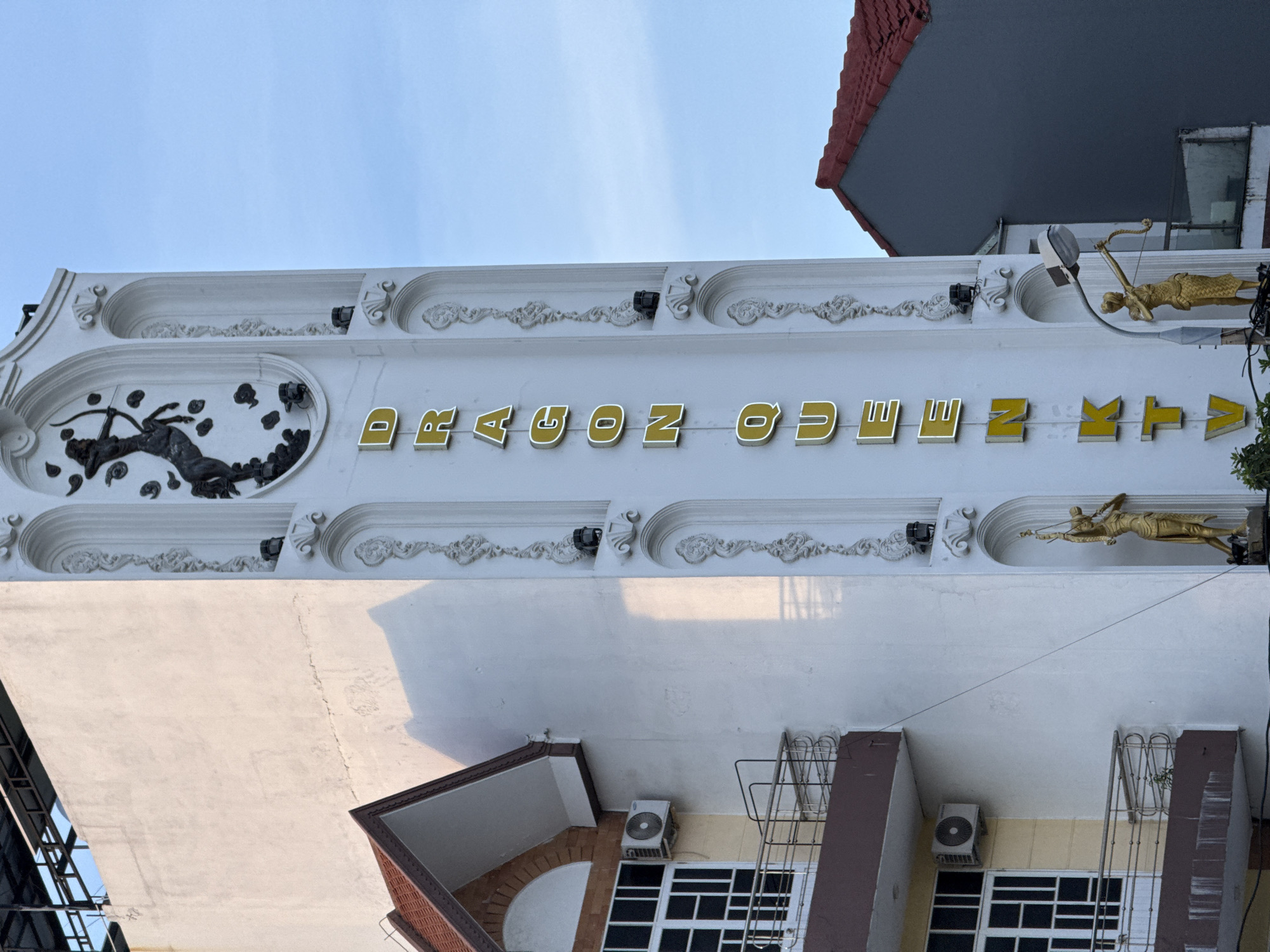 This is an outdoor, eye-level, medium shot of the upper facade of a building in Đà Nẵng, Vietnam, on a clear day. The building's white exterior is decorated with ornate relief carvings and golden statues. Prominently displayed across the facade is the name "DRAGON QUEEN" in large, golden, 3D letters with white outlining. To the left of the text, within an oval relief, is a dark sculpture of what appears to be a deer or similar animal in mid-leap. To the right of the text, two golden statues of figures holding bows and arrows are mounted on the facade. The sky above is a pale blue with no clouds. Below the ornate upper level, the building has multiple windows and air conditioning units visible. The overall impression is one of an older building with elaborate architectural details.