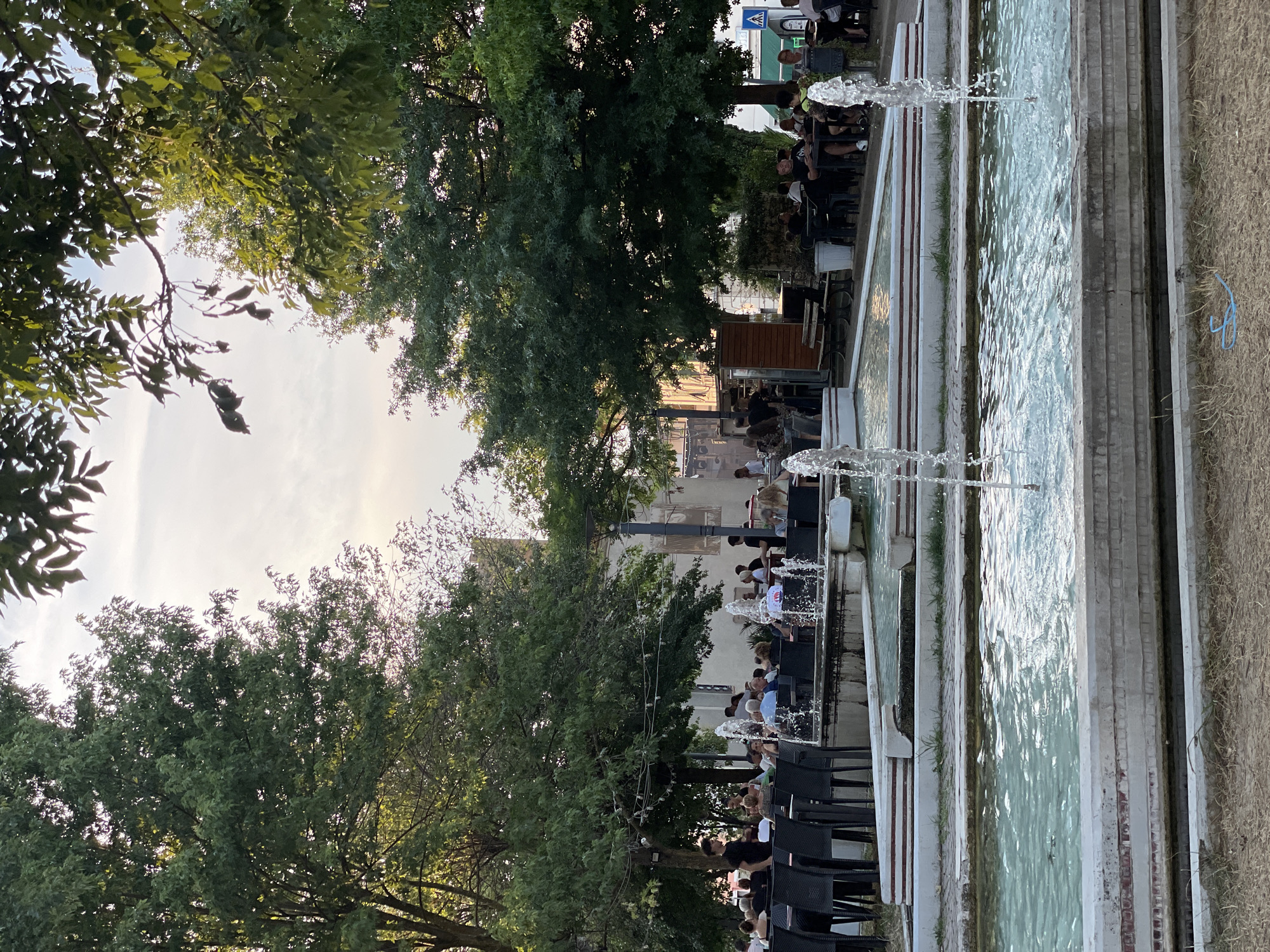 In Altopascio, Italy, an outdoor seating area surrounds a tiered fountain. The fountain features multiple levels of water jets. On the left side of the image, black chairs are arranged around tables, with people seated. A server in a black shirt stands near the tables. A large tree with dense green foliage provides shade. Further back, additional tables and chairs are visible, occupied by patrons enjoying the outdoor ambiance. A building with white walls and a dark roof stands in the background, partially obscured by trees. The sky above is bright with some clouds. The foreground shows dry grass and a blue plastic object.