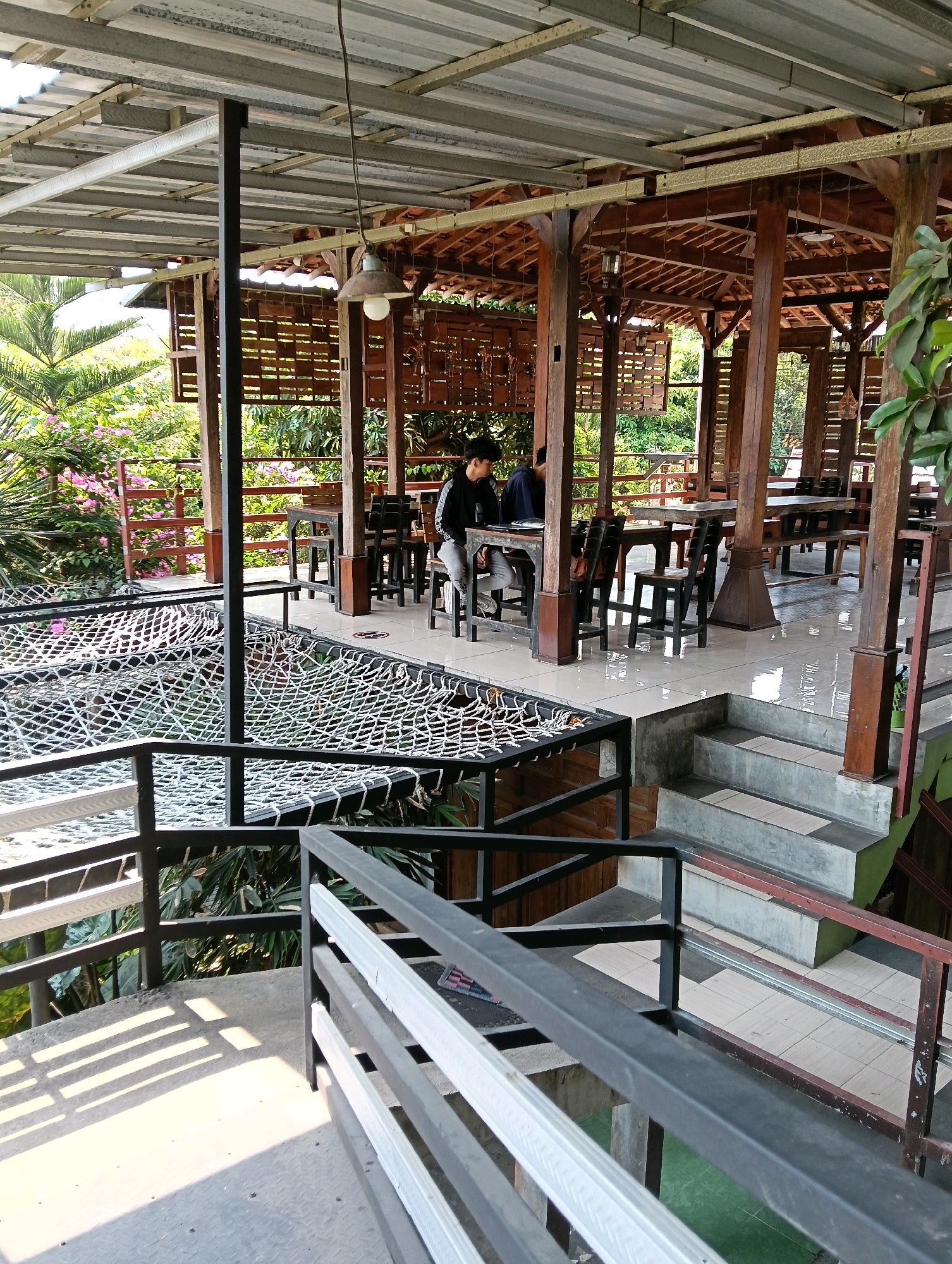 The image depicts an open-air seating area at a restaurant or cafe, possibly located in Papahan, Indonesia. In the foreground, a metal railing runs horizontally across the frame, with a concrete floor below. Beyond the railing, a netted area, possibly a hammock-like seating arrangement, is visible. A set of concrete stairs leads down from the upper level to a lower level. The middle ground features a covered dining area with wooden pillars and a corrugated metal roof supported by a metal frame. Several wooden tables and chairs are arranged for diners. Two individuals are seated at one of the tables, one looking down at a laptop or notebook and the other facing away from the camera. In the background, lush green foliage and pink flowers are visible, suggesting an outdoor or garden setting. The overall atmosphere appears relaxed and informal.