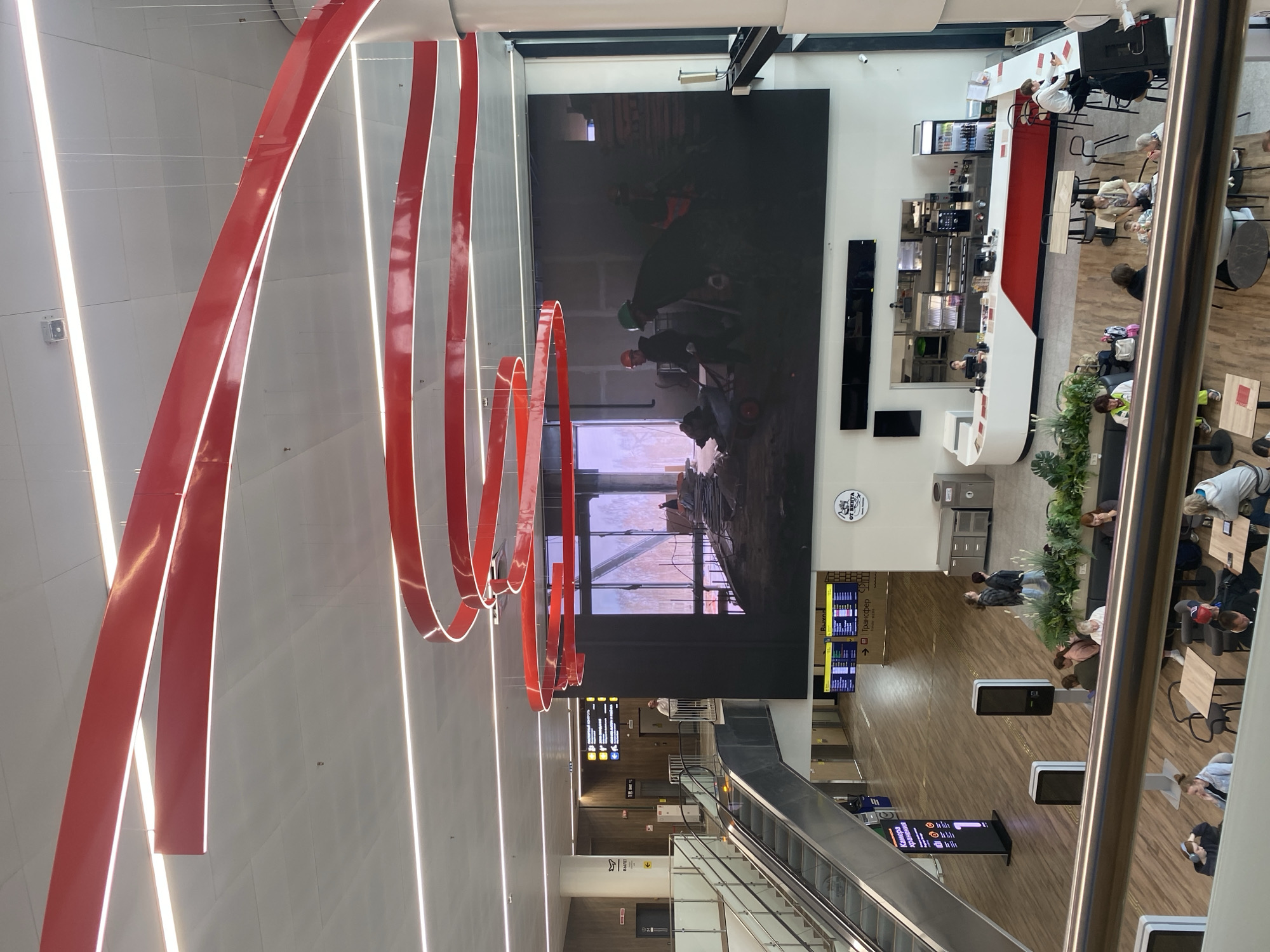 The image depicts the interior of a modern airport or public transport hub in Новое Мартьяново, Russia. The scene is characterized by high ceilings, ample natural light, and contemporary architecture. Red, curved lighting fixtures with integrated white LED strips are suspended from the ceiling, creating a striking visual element.
Dominating the central area is a massive digital screen displaying a construction site, showing workers in hard hats and safety vests operating machinery and moving materials. To the left, an escalator ascends alongside a large pillar, with directional signage for "ВЫХОД" (Exit) and " КАМЕРА хранения" (Storage Room) visible. Further in the background, digital departure boards display flight or train information.
The ground floor features a cafe or food service area with a white and red counter, behind which staff are working. Tables and chairs are arranged in a seating area, with some patrons visible. To the right of the cafe, a bar-style counter with high stools offers additional seating, and a person is seated there.
In the foreground, a metal railing separates the viewer's vantage point from the rest of the space, suggesting the photo was taken from an upper level or balcony. Several people are seated at tables in this foreground area, some engaged with their phones or laptops, others in conversation. Lush green plants are strategically placed throughout the space, adding a touch of nature to the environment. The overall atmosphere appears to be one of transit and waiting.