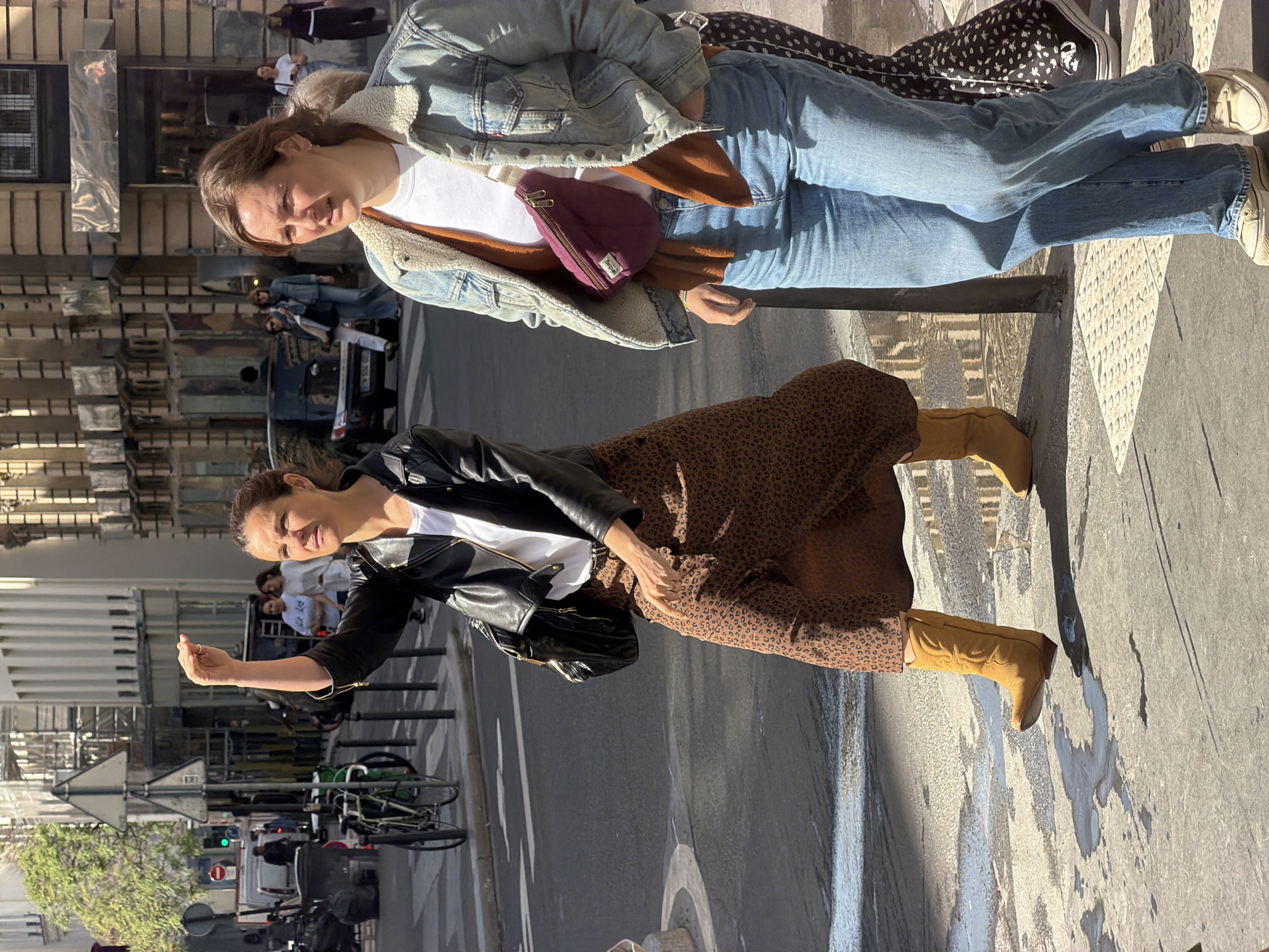 Two women stand together on a sidewalk in Paris, France. The woman on the left wears a black leather jacket, a white t-shirt, a brown patterned skirt, and brown boots. She holds her hand up as if she's calling a taxi or someone's attention. The woman on the right wears a light blue denim jacket with a shearling collar, a white t-shirt, blue jeans, and white sneakers. She also has a small maroon bag slung across her chest. In the background, there is a street with cars, bicycles, and pedestrians. Buildings and street signs typical of Paris are also visible.