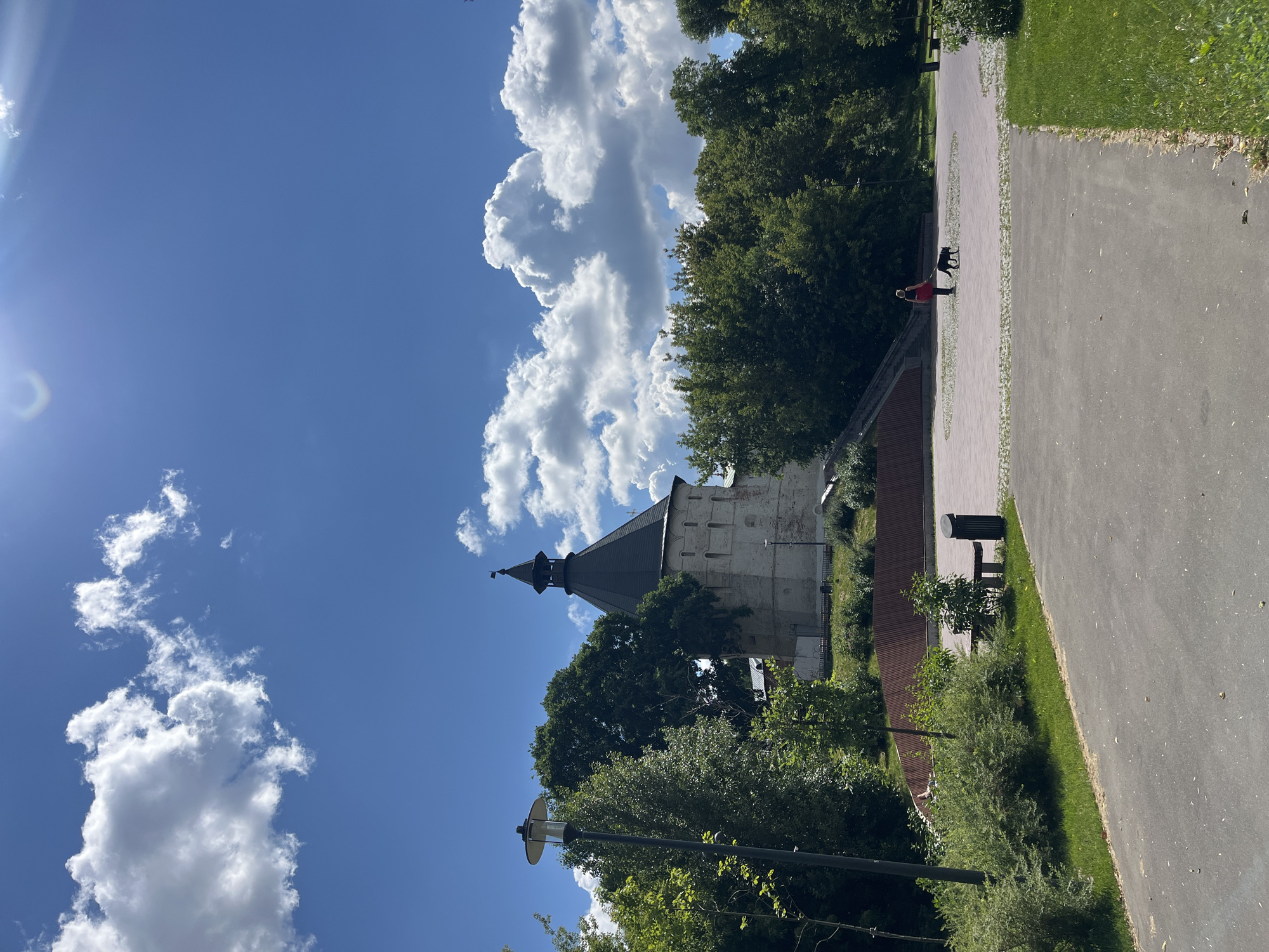 This media file, taken in Moscow, Russia, depicts a sunny day in a park with a historic structure visible. The scene is oriented vertically, with the sky occupying the upper portion of the frame and the ground below.
The sky is a vibrant blue, dotted with fluffy white cumulus clouds, suggesting fair weather. A lens flare from the sun is visible in the upper left corner, indicating it's likely daytime, possibly late morning or early afternoon.
In the middle ground, a weathered, stone building with a dark, conical roof stands amongst lush green trees. This building appears to be a tower or part of a larger historical complex. A person is walking a black dog on a paved pathway that runs through the park. The pathway is bordered by green grass and more trees. There is a trash can and a bench visible near the pathway.
The overall mood is peaceful and pleasant, characteristic of a summer day spent outdoors. The historical architecture, combined with the natural elements of the park and the presence of people and a pet, creates a sense of a serene urban escape. No visible text is present in the image.