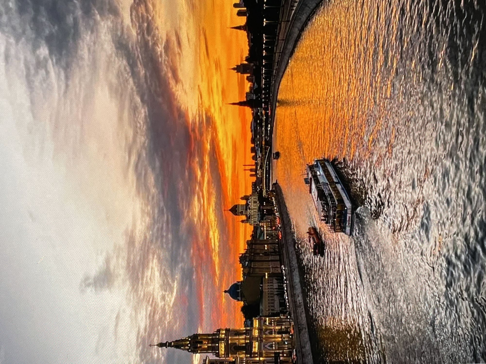 This is a vertical, eye-level, medium shot of a sunset over the Moskva River in Moscow, Russia. The sky is a dramatic mix of orange, yellow, and gray clouds, with the sun's light reflecting intensely on the water. Buildings line the riverbank, including a tall, ornate tower on the left, and other structures with domes and spires. Several boats are visible on the river, including a large white passenger boat in the foreground and a smaller red boat behind it. The water is choppy, with reflections of the sky and buildings visible. The scene appears to be illuminated by the sunset and artificial lights from the buildings.