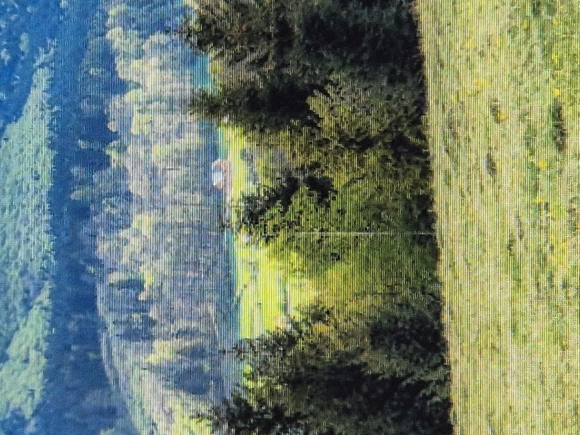 The image depicts a pastoral scene in Sozonivka, Ukraine. In the foreground, a green field with yellow wildflowers stretches towards a dense forest. The forest forms a dark green band across the middle ground, behind which a bright green meadow is visible. Nestled within the meadow, a small, red-roofed building with white walls stands out. Further in the background, a distant blue mountain range is silhouetted against a hazy sky. The image has a slightly grainy texture, with horizontal lines running through it.