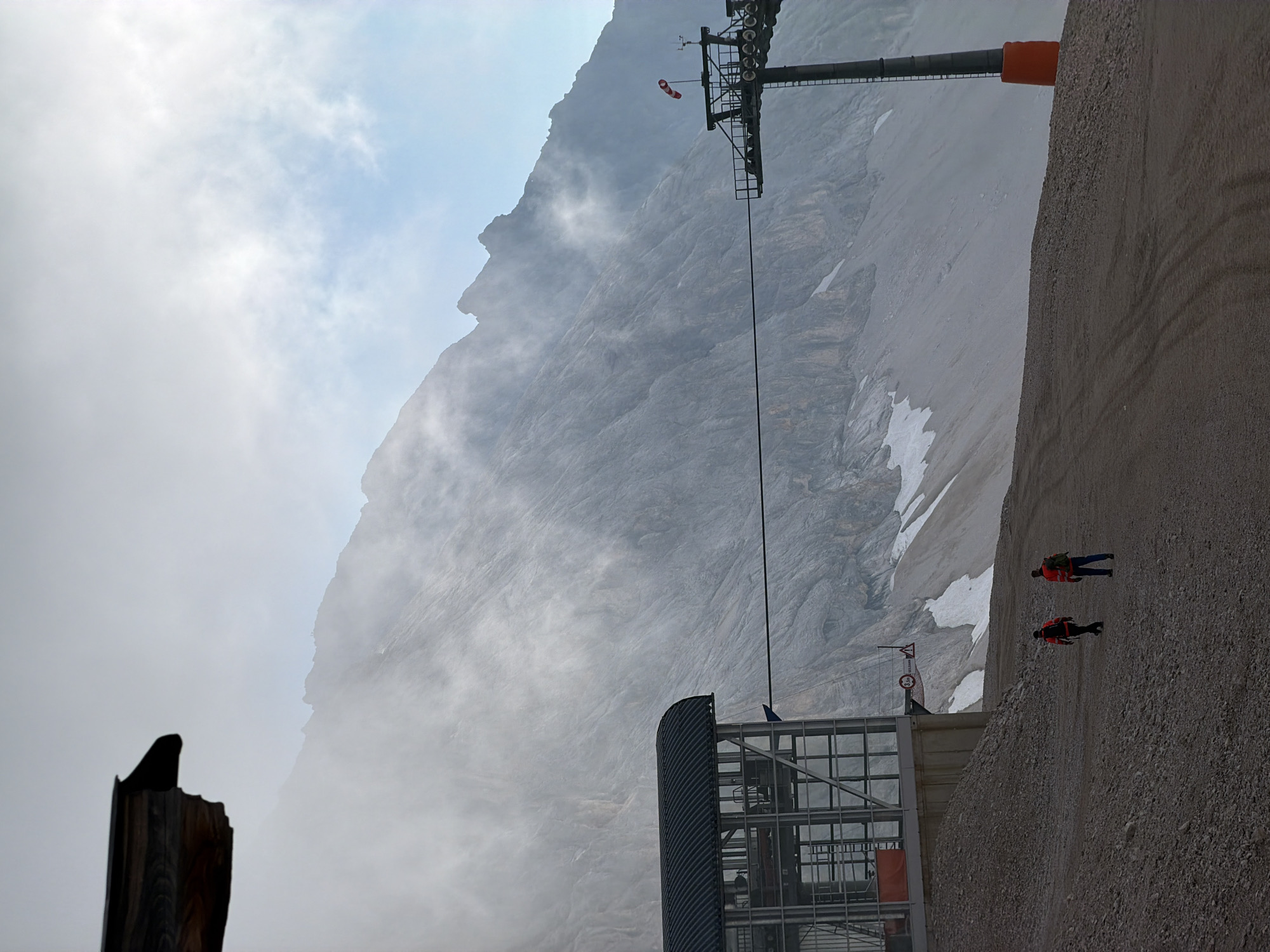 This image depicts a high-altitude scene on the Zugspitze, Germany's highest peak, near Garmisch-Partenkirchen. In the foreground, a gravel path with tire tracks leads towards a modern building with a dark, curved roof and glass walls. Two individuals wearing bright orange safety vests walk away from the viewer on the path. To the right, a ski lift tower stands tall, topped with a windsock and what appears to be meteorological equipment. The tower has a bright orange protective sleeve at its base. A thick cable extends from the ski lift structure across the image. Behind these elements, a massive, rugged mountain face dominates the background, partially obscured by mist and clouds. Patches of snow are visible on the mountain slopes. The sky above is a blend of light gray clouds and patches of blue. The overall impression is one of an industrial infrastructure set against a dramatic, natural mountain environment.