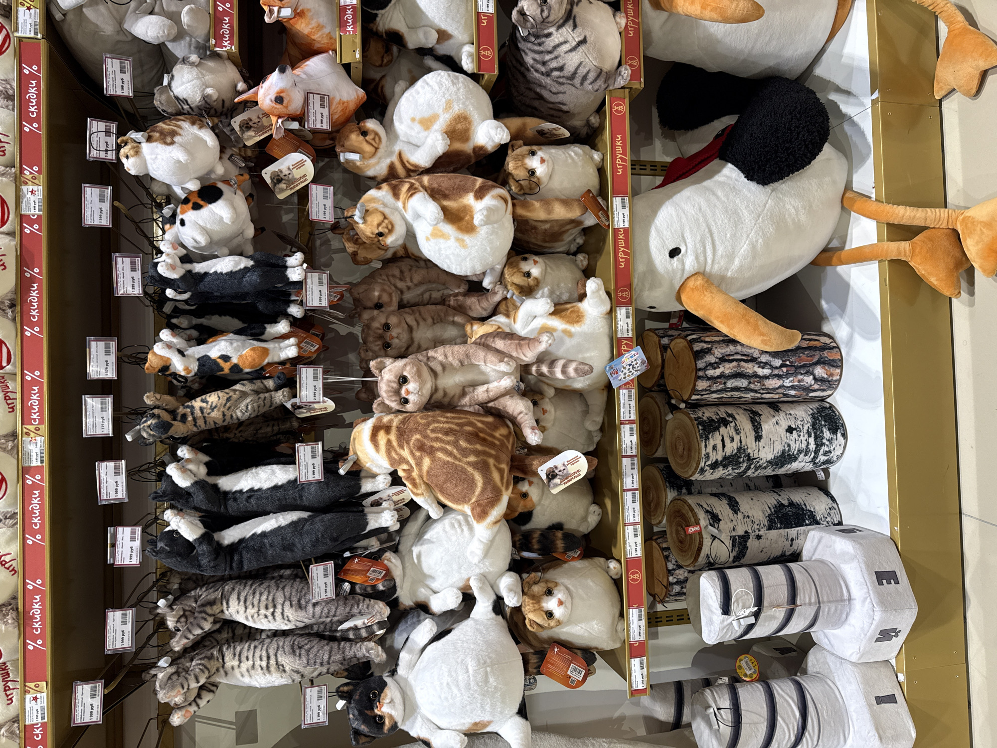 The image depicts a shelf in a store, presumably in Moscow, Russia, given the Cyrillic characters on the promotional strips. The shelf is densely packed with plush toys. The left side of the shelf predominantly features plush cat toys of various colors and patterns. To the right, there are plush logs and a large plush white duck with a black head and orange beak and feet. Price tags are visible on the toys. The scene is brightly lit and gives the impression of a retail setting.