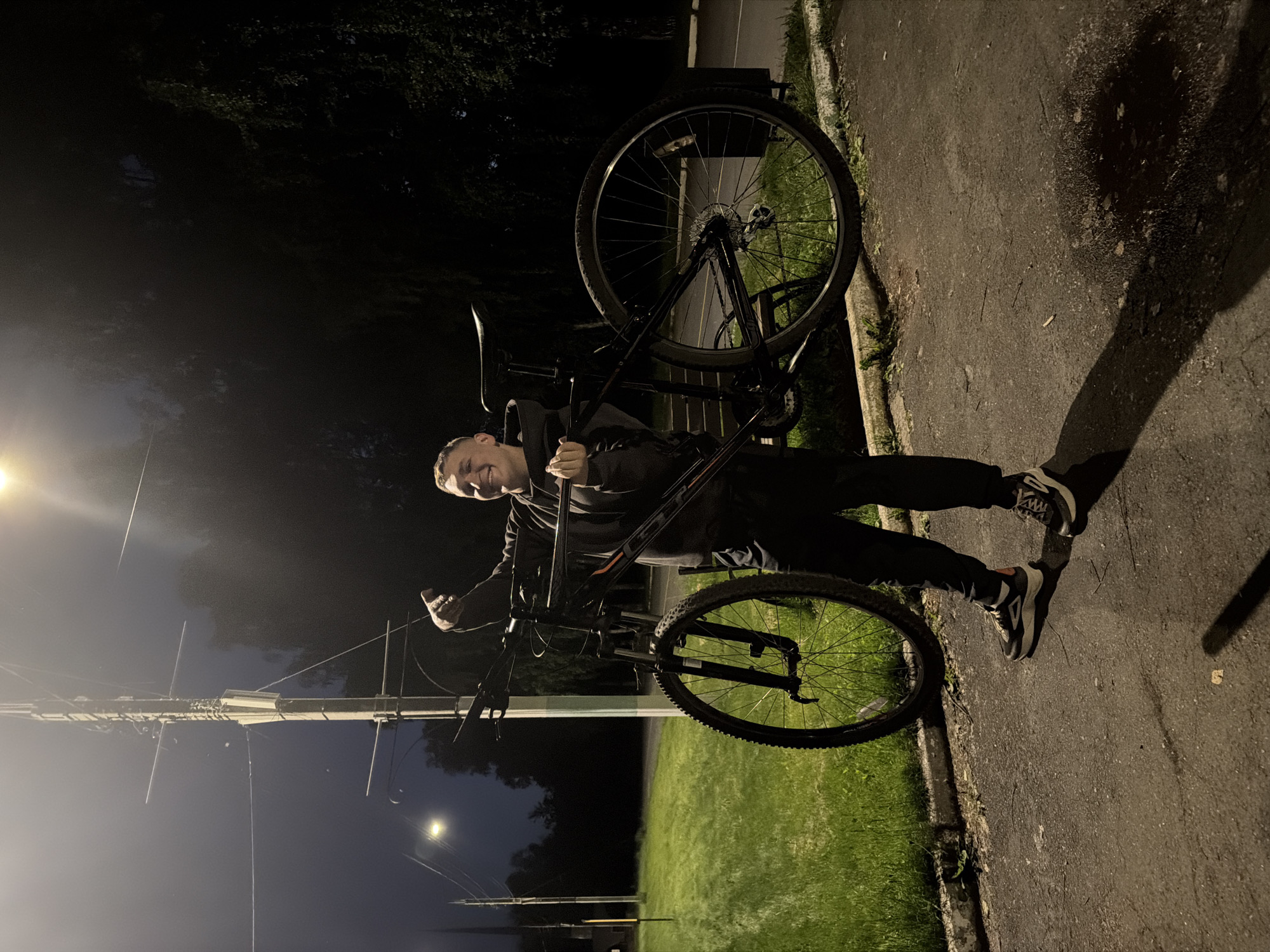 The image, taken at night in Izhevsk, Russia, features a young man holding a mountain bike on an asphalt path. The man, who appears to be in his late teens or early twenties, is smiling and looking directly at the camera. He is dressed casually in a dark hooded sweatshirt, dark pants, and athletic shoes.
He is holding a black mountain bike with subtle orange accents on its frame; the brand "GT" is visible on the bike's downtube. The bike's front wheel is lifted slightly off the ground, as he holds it by the frame near the handlebars.
The scene is illuminated by a strong artificial light source, likely a streetlight, positioned to the left of the frame. This light casts long, distinct shadows of the man and his bicycle onto the asphalt path and the adjacent grassy area. In the background, dark trees are visible, along with another distant streetlight. A utility pole with overhead wires is also present on the left side of the image. The ground consists of a clear asphalt path bordered by a concrete curb and a grassy verge. The man's cheerful expression suggests a positive mood.