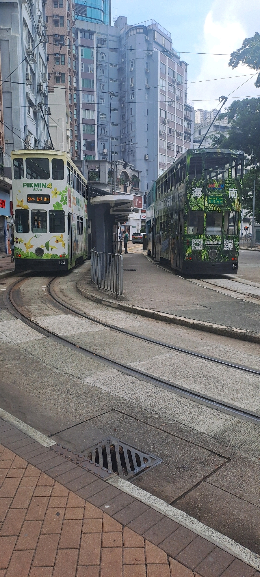 In Hong Kong, China, two green double-decker trams are positioned on tram tracks on a street. The tram on the left is white with green accents and features characters from the game Pikmin 4. The tram on the right is predominantly green and decorated with jungle-themed imagery. Both trams have advertising on their sides. The scene is set against a backdrop of tall, densely packed apartment buildings, characteristic of Hong Kong's urban landscape. A tram stop shelter is visible between the two trams, with a person standing near it. The street surface is concrete, and a drainage grate is present in the foreground. The sky is overcast.