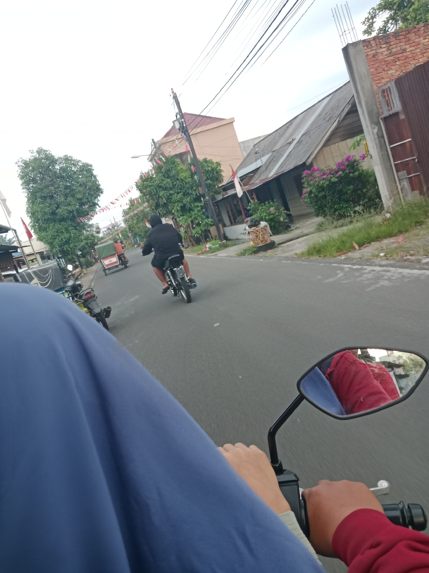 In the city of Binjai, Indonesia, a person is riding a motorcycle on a paved road. The perspective is from the rider's point of view, showing their hands on the handlebars and a blue garment in the foreground. In the distance, another motorcyclist wearing a black shirt is riding ahead, followed by a three-wheeled vehicle. The road is lined with trees, houses, and a brick wall. The sky is overcast. A motorcycle mirror reflects the red garment of the rider.
