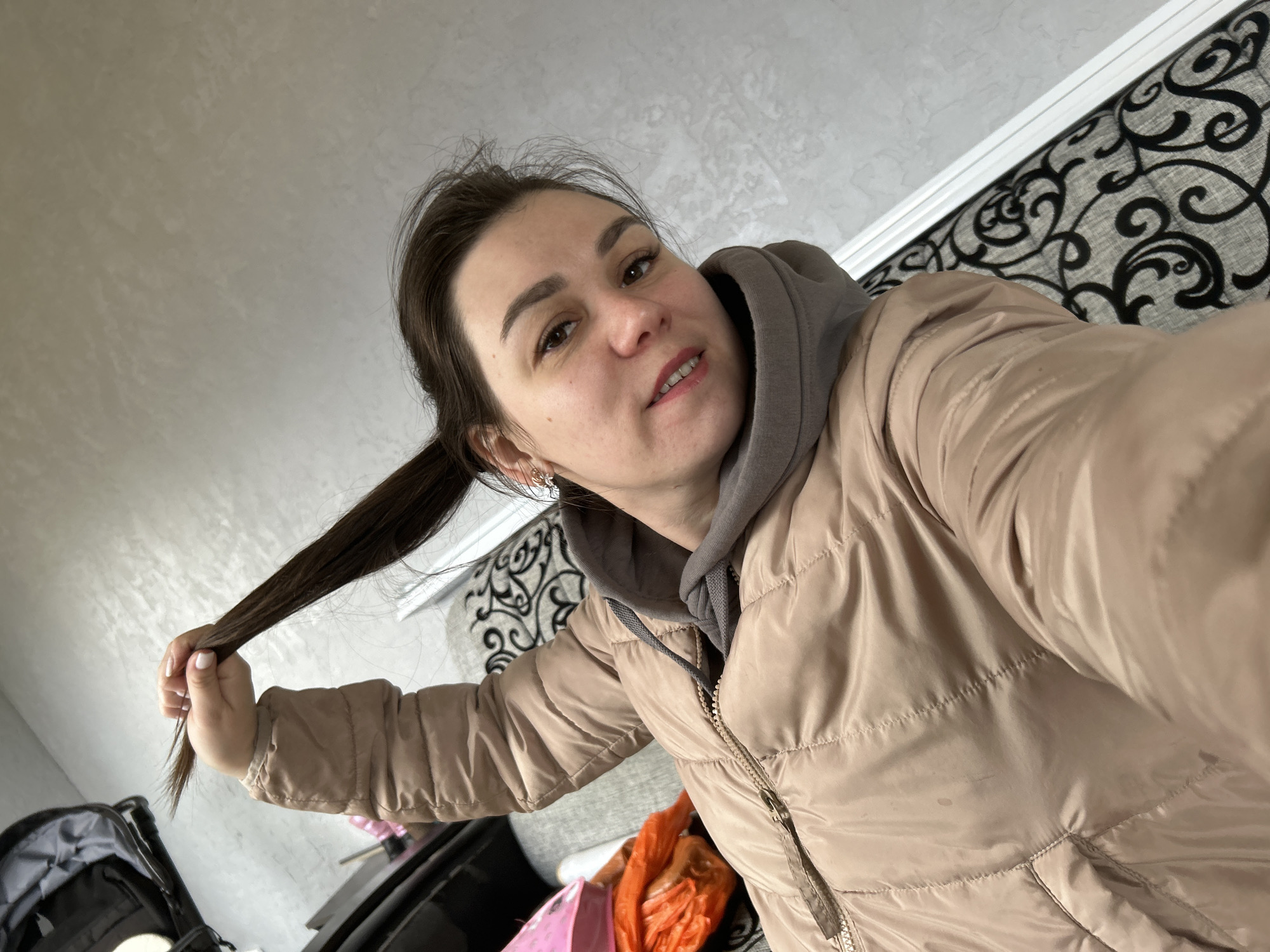 A selfie image shows a woman indoors, holding a section of her dark brown hair in her left hand. She is wearing a beige puffer jacket over a gray hooded sweatshirt. The background includes a gray wall, a patterned headboard or wall covering in black and white, and glimpses of objects like a black suitcase or bag. The location is likely indoors in an unidentified building, possibly in the city of Chaikovsky, Russia, but there are no visual cues to confirm this specific location.