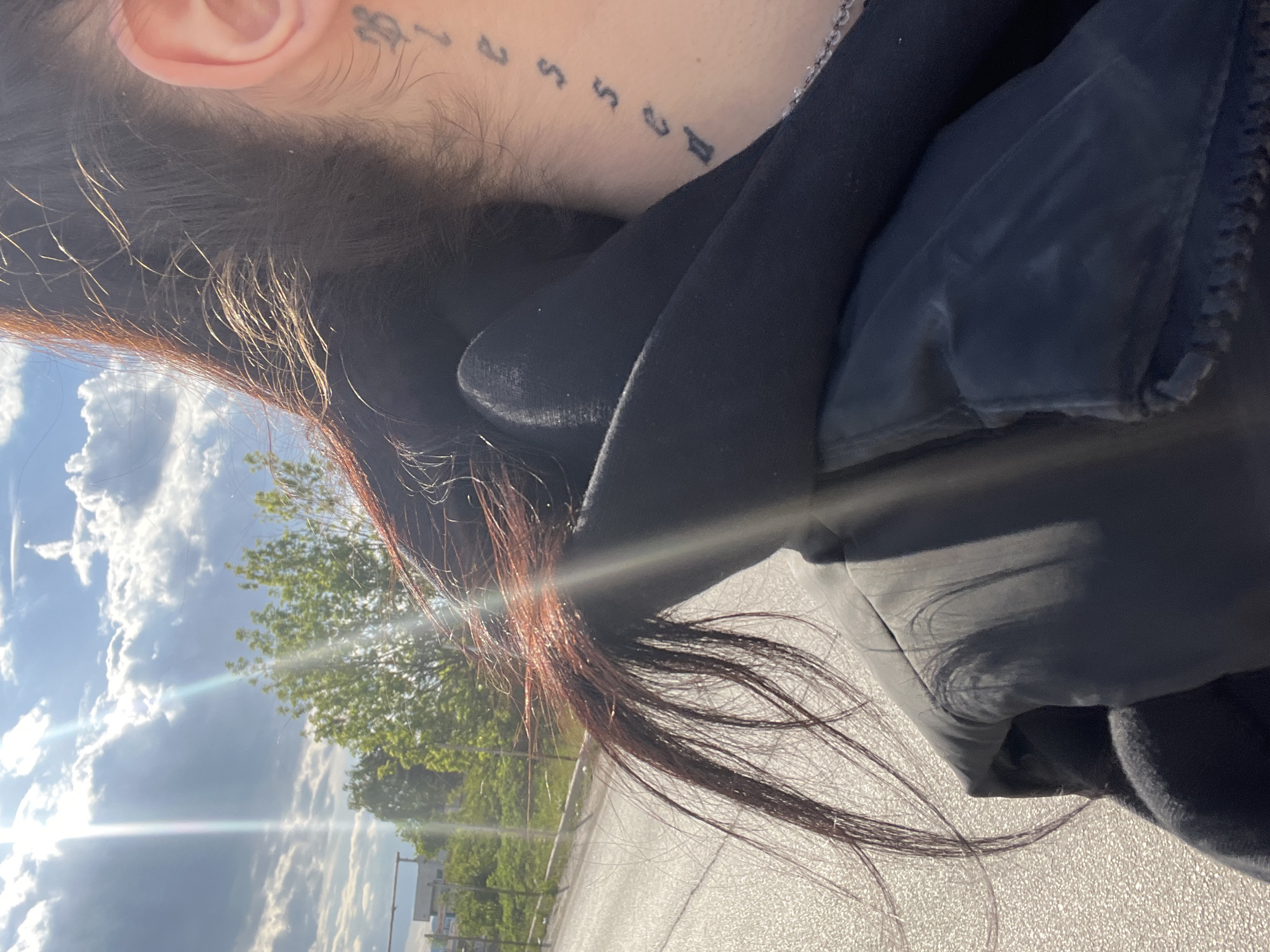 A medium shot captures a subject outdoors. The subject has brown hair with red highlights, and a tattoo of numbers on the neck near the ear. The person is wearing a black jacket. The background shows a landscape under a cloudy sky with trees. The location appears to be a city street, potentially in Sofia, Bulgaria. The image is slightly rotated.