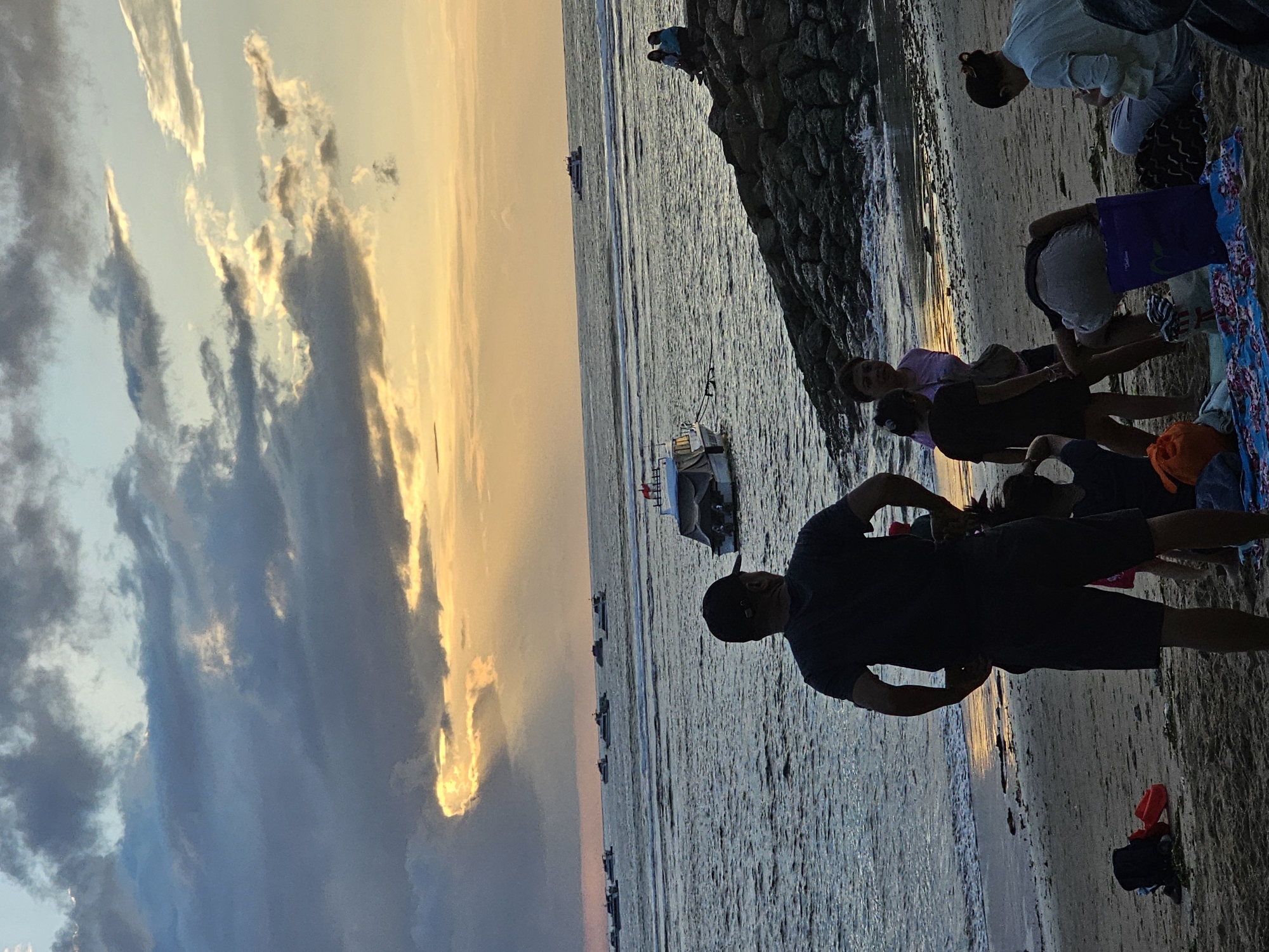A group of people are on a sandy beach in Denpasar, Indonesia, near a rocky breakwater. The sun is setting, casting a warm glow over the ocean and the sky, which is filled with dramatic clouds. Several boats are visible on the water.
In the foreground, a man wearing a dark t-shirt and a cap stands with his back to the camera, looking out at the sea. To his right, a woman in a pink shirt and two children are on the sand. One child, wearing a dark t-shirt and shorts, is bending over. Another person in a light blue shirt is sitting on the sand.
To the right of the frame, two individuals are seated on the rocky breakwater, also facing the ocean. In the background, multiple boats are scattered across the water. The tide appears to be going out, as evidenced by the wet sand and some debris on the beach. The overall atmosphere is peaceful and serene, suggesting a typical evening scene at a popular beach location.