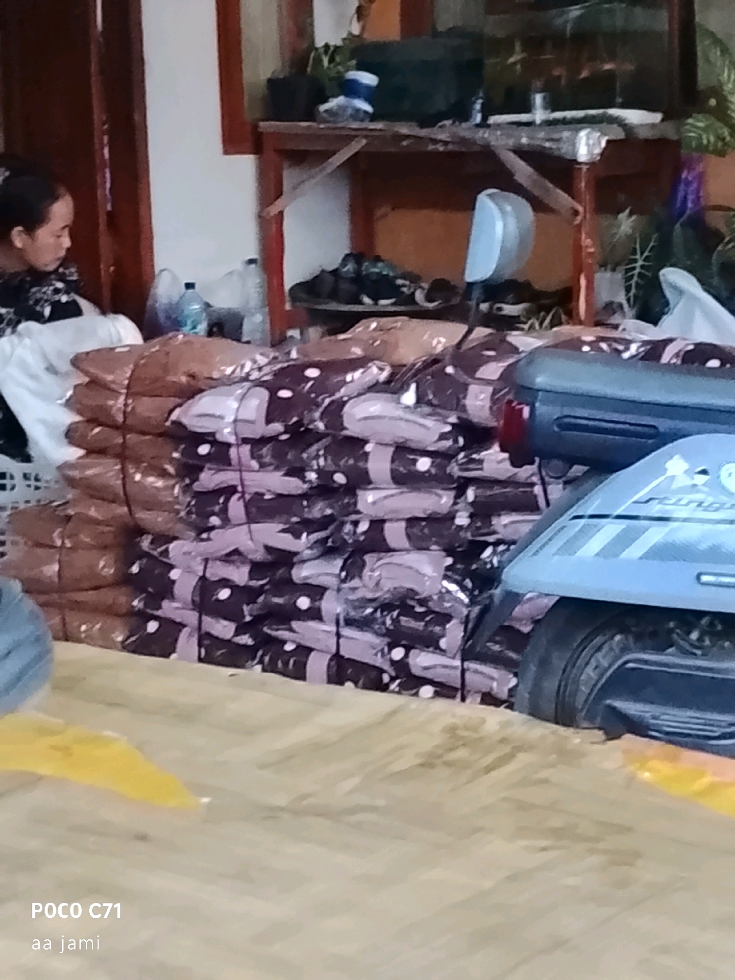 The image depicts a close-up view of stacks of packaged goods, likely blankets or textiles, in what appears to be a storage or sales area. The packages are predominantly in dark brown and purple hues with a white polka dot pattern, neatly stacked and bound with dark string. In the background, a woman with dark hair tied back is partially visible, looking down. A shelf unit with various items, including bottles and shoes, is also present. A portion of a light blue vehicle, possibly a scooter or motorcycle, is in the foreground on the right side of the image, with its tail light visible. The overall environment suggests an indoor setting, possibly a shop or warehouse. There is text overlay on the bottom left corner of the image that reads "POCO C71" and "aa jami", which are likely camera model and a username or tag. The scene appears to be located in Tasikmalaya, Indonesia, based on the provided context, although no specific visual cues within the image confirm this location.