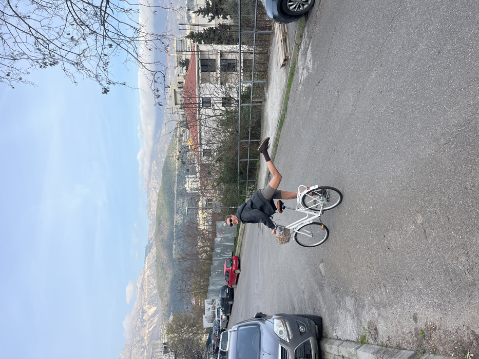 The image displays an outdoor daytime scene on an asphalt road in a semi-urban area of Budva, Montenegro, with mountainous terrain in the background.
A single individual, appearing female, is positioned on a white city-style bicycle in the center-right of the frame. She wears a dark grey hooded jacket, dark sunglasses, light brown shorts, dark brown socks, and dark brown shoes. Her right leg is extended horizontally backward, bent at the knee, with the foot raised, while her left foot remains on the ground. Her head is turned towards the camera, and she is smiling. Her hands rest on the bicycle handlebars. The white bicycle features a brown seat and handlebars, a front-mounted woven basket containing light-colored material, a rear rack, and a red reflector.
To the left of the road, a silver-grey Ford Kuga SUV is parked alongside a concrete curb, facing away from the camera. Additional parked vehicles, including a red car, are visible further down the street behind the Ford. Temporary construction hoarding or fencing is present behind these parked vehicles.
On the right side of the road, a low metal guardrail separates the pavement from an overgrown area containing bare deciduous trees and shrubs, indicative of a non-summer season. Several white wooden pallets are visible on the ground behind the guardrail. Beyond this vegetation, residential buildings with white facades and distinctive red tiled roofs are visible.
The background is dominated by a range of large, greyish-brown mountains, with some peaks partially obscured by low-lying white clouds. A clear blue sky is visible above the mountains.