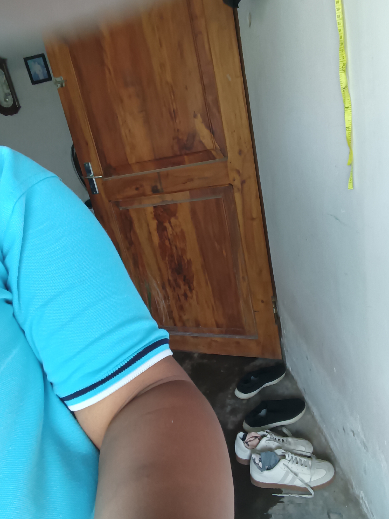 In Krampilan, Indonesia, a person wearing a blue polo shirt stands near a wooden door. Shoes are scattered on the floor: two black slip-on shoes and two pairs of white sneakers. A yellow measuring tape hangs vertically on a white wall.