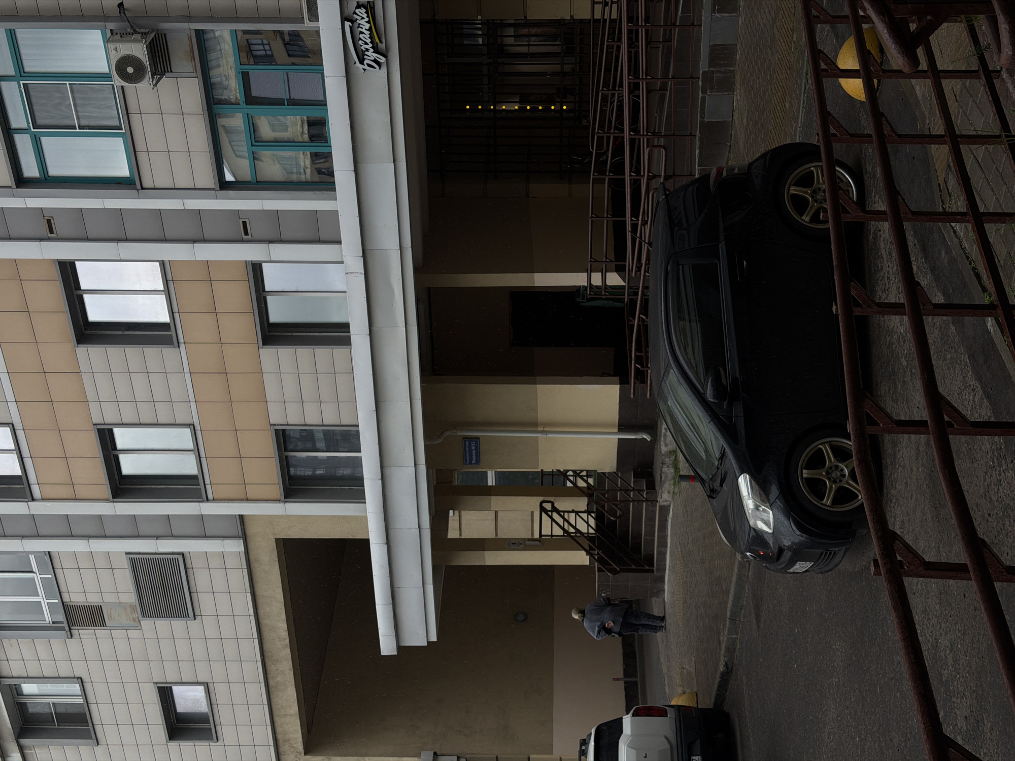 The image depicts an exterior scene of a modern building with a tiled facade and numerous windows. The building's architecture is characterized by a mix of beige and grey tiles, with some windows featuring teal frames. An air conditioning unit is visible mounted on the building. Adjacent to the building is a dark-colored car, possibly a hatchback, with gold-colored rims. A metal structure, resembling a fire escape or scaffolding, runs vertically along the right side of the image.
The scene appears to be an urban setting, with a paved ground surface. In the background, there are stairs leading up to an entrance, and a person, dressed in a grey jacket and dark pants, stands near the bottom of the stairs. A street sign with Cyrillic text is mounted on the wall. Further down, a white car is partially visible.
The lighting suggests it might be daytime, but the overall mood is somewhat subdued, possibly indicating overcast weather or a gloomy atmosphere. The presence of rain droplets on the ground and car suggests it is raining. The text visible on the building facade reads "Буханка," which translates to "Loaf" and appears to be a business name. The street sign also contains Cyrillic text, though it is not entirely legible. The setting is likely a residential or commercial area in Moscow, Russia.