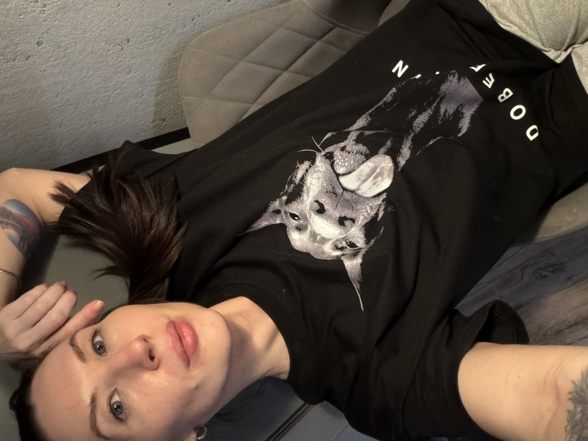 A fair-skinned person with shoulder-length dark brown hair is lying down in what appears to be an indoor setting, possibly in Perm, Russia. The person is wearing a black t-shirt with a graphic of a Doberman dog on it. The person's head is tilted towards the camera, and their face is visible, showing blue eyes, full lips, and light makeup. The person's left hand is raised towards their face, and there is a tattoo visible on their left forearm. A grey upholstered chair is behind the person. A wall and a wood-look floor are also visible.