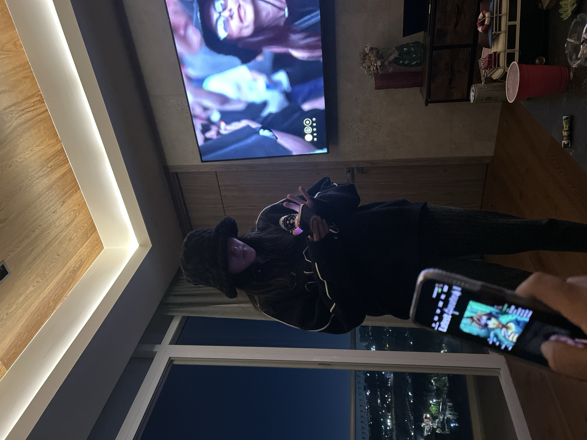 A person in a dark furry hat and black jacket stands in front of a large window overlooking a night cityscape in Huixquilucan de Degollado, Mexico. They are holding something in their hands, possibly a phone or other small device. In the foreground, another person is holding a phone, with the screen displaying an image. Behind the person in the hat is a large television screen showing what appears to be a concert or performance. To the right of the television, there is a shelf with various items, including a red cup and a can.
