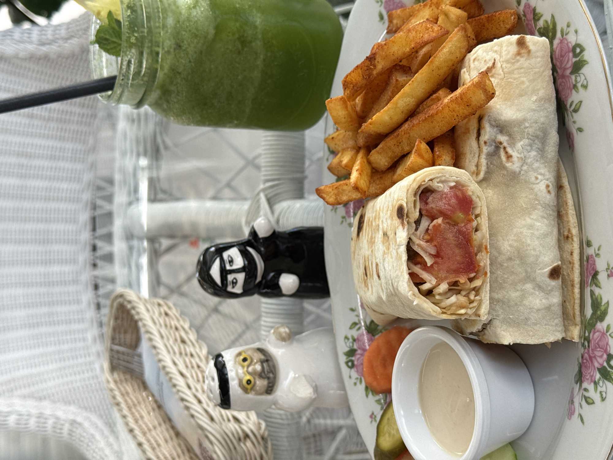 The image, presented vertically, captures a bright and inviting meal setting, likely outdoors or in a well-lit indoor space.
A tall glass jar filled with a frothy, vibrant green beverage, possibly a mint lemonade or smoothie, is positioned on the upper left. It features a black straw and is garnished with mint leaves and a citrus wedge. To the right, a white oval plate with an elegant pink floral rim holds the main course. A generous portion of golden, seasoned french fries is piled high, alongside two wraps, resembling shawarmas. One wrap is partially unwrapped, revealing a filling of sliced tomatoes and what appears to be shredded cabbage or lettuce. A small white ramekin containing a light, creamy dipping sauce (such as tahini) is also on the plate, accompanied by slices of carrots and pickles.
Between the drink and the plate, two distinctive small figurines add a cultural touch. One, in black, depicts a figure in traditional abaya or niqab. The other, in white, portrays a figure in traditional Arab male attire, including a ghutra and glasses, with a slightly caricatured, serious expression. The meal is presented on a textured white surface, possibly a wicker table or a patterned tablecloth. A small, light-colored woven basket is partially visible in the lower left.
The bright illumination suggests a daytime scene. The specific food items and the cultural attire represented by the figurines strongly indicate a Middle Eastern culinary theme, consistent with a specialized restaurant, even given the context of Podgorica, Montenegro. No visible text is present in the image.