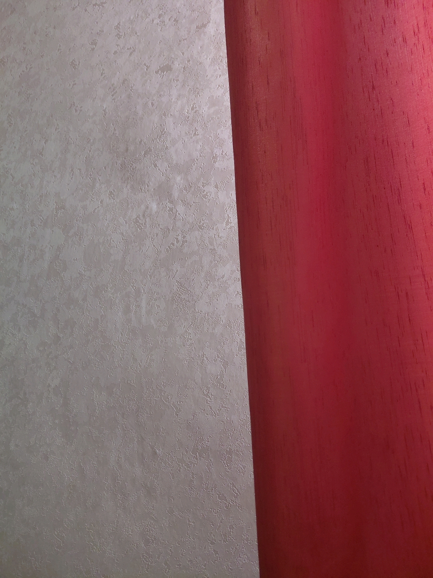 The image shows a close-up view of a textured, light-colored wall on the left, and a vibrant red fabric on the right. The fabric appears to be a curtain or drapery, with visible vertical threads and a slightly uneven texture. The light wall has an irregular, mottled pattern that gives it depth. The lighting suggests the image was taken indoors, with a light source casting subtle shadows. There are no people or any discernible features that would indicate a specific location in Malinovka, Russia. The scene is static and focuses solely on the textures and colors of the two distinct surfaces.