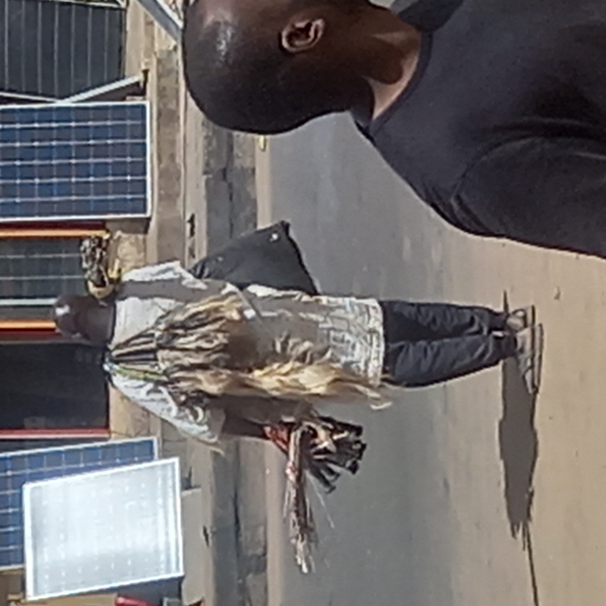 A man dressed in traditional garb with a long, flowing skirt and carrying an object, possibly ceremonial, walks along a street in Sardauna, Nigeria. Solar panels are visible on a building in the background. Another individual in a dark shirt is partially visible in the foreground. The environment suggests an urban setting in Sardauna, Nigeria.